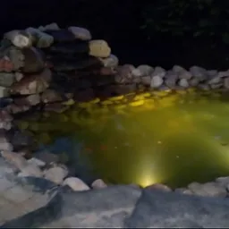 Oświetlony oczko wodne z kamienną kaskadą i obrzeżem z kostki, widoczne w nocy.