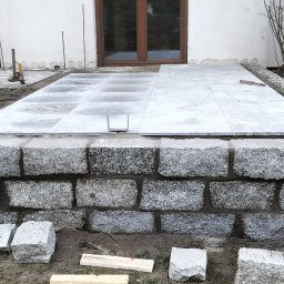 BrukPol Wiesław Kraiński - Wykonywanie tarasu z kamiennych płyt przy białym budynku, widoczne kamienne obrzeże, płyty tarasowe i narzędzia, etap prac w toku.