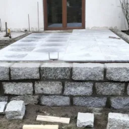 Wykonywanie tarasu z kamiennych płyt przy białym budynku, widoczne kamienne obrzeże, płyty tarasowe i narzędzia, etap prac w toku.