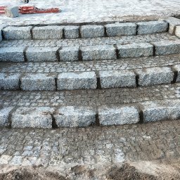 BrukPol Wiesław Kraiński - Schody z kostki granitowej w trakcie budowy, widoczne narzędzia i materiały budowlane, trawa w tle.