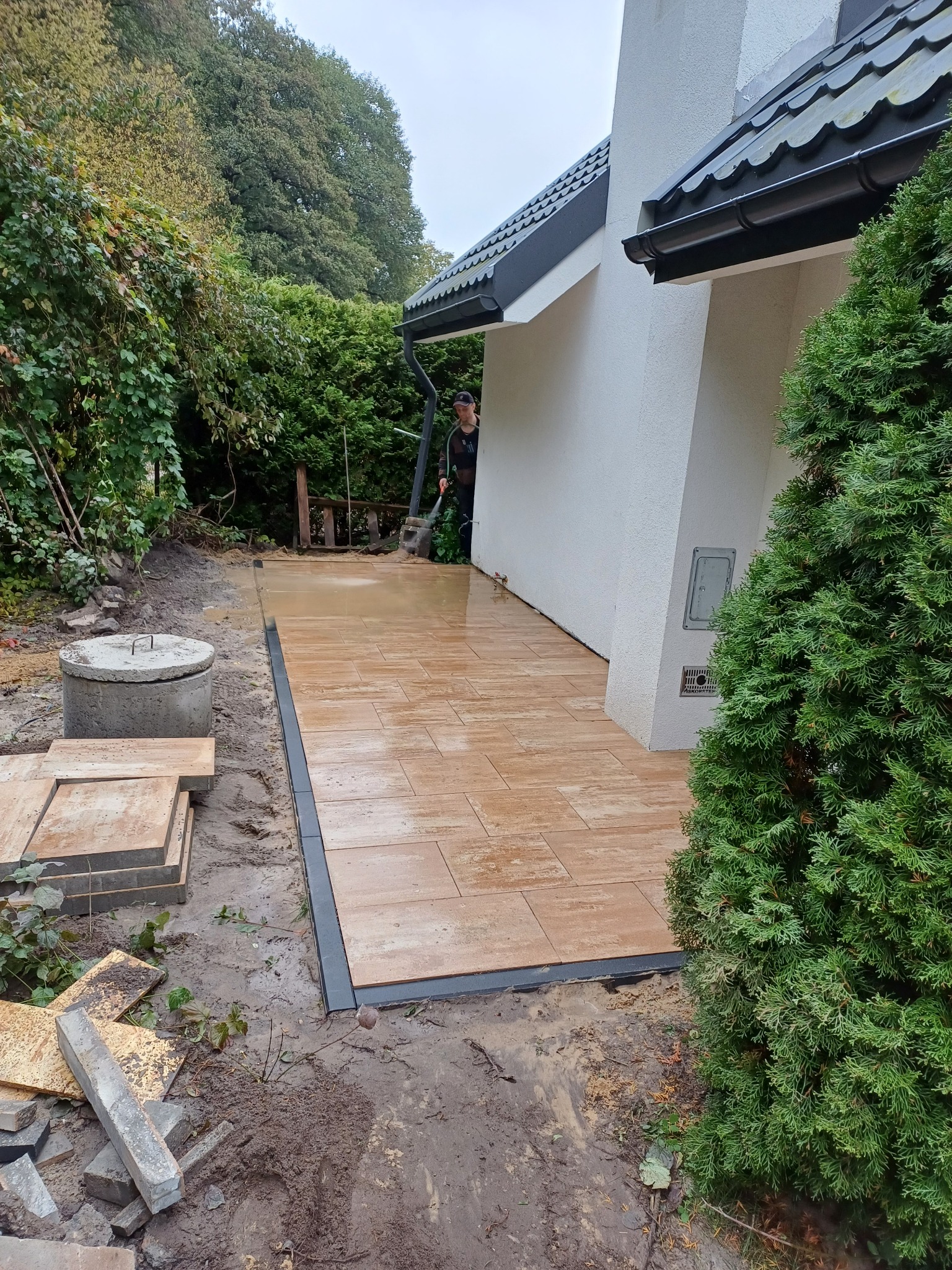 Umyta, brązowa powierzchnia z prostokątnych płyt na zewnątrz budynku, obok trawnika z drzewami i krzewami, z widocznym pracownikiem spłukującym resztki brudu.