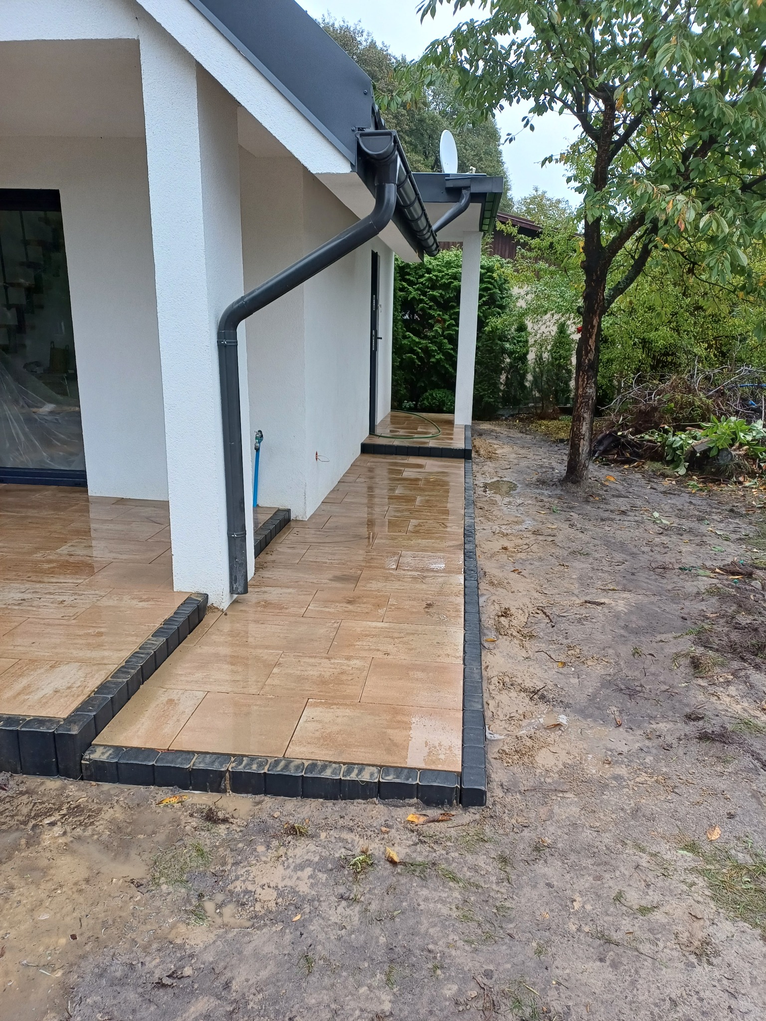 Nowoczesny taras przydomowy z jasnych płyt, obramowany ciemną kostką, po deszczu, widok na biały budynek z czarnymi rynnami i zielenią w tle.