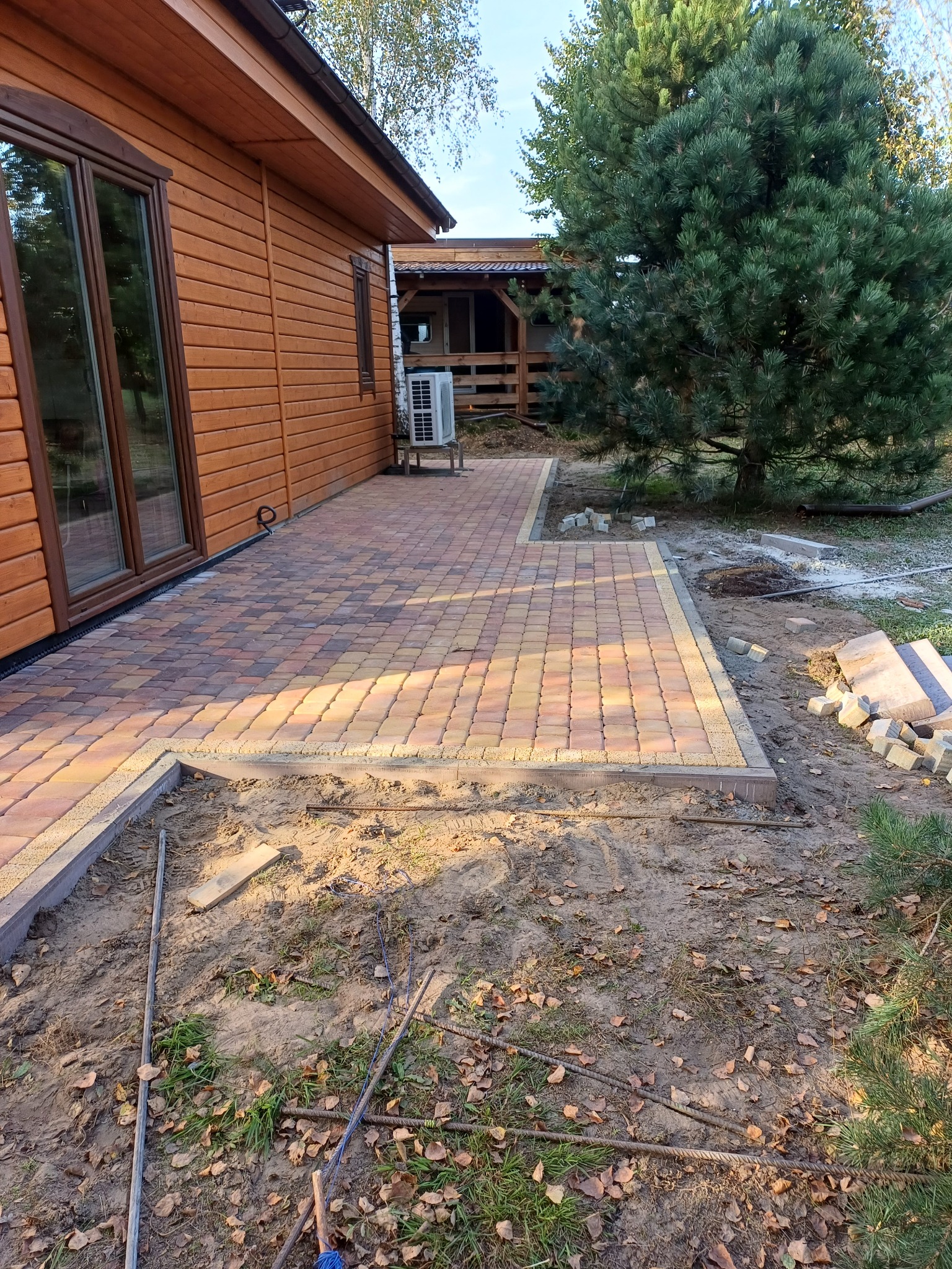 Fragment układanej nawierzchni z kostki brukowej w odcieniach brązu i żółci przy drewnianym domu, widoczne narzędzia i przygotowane podłoże.