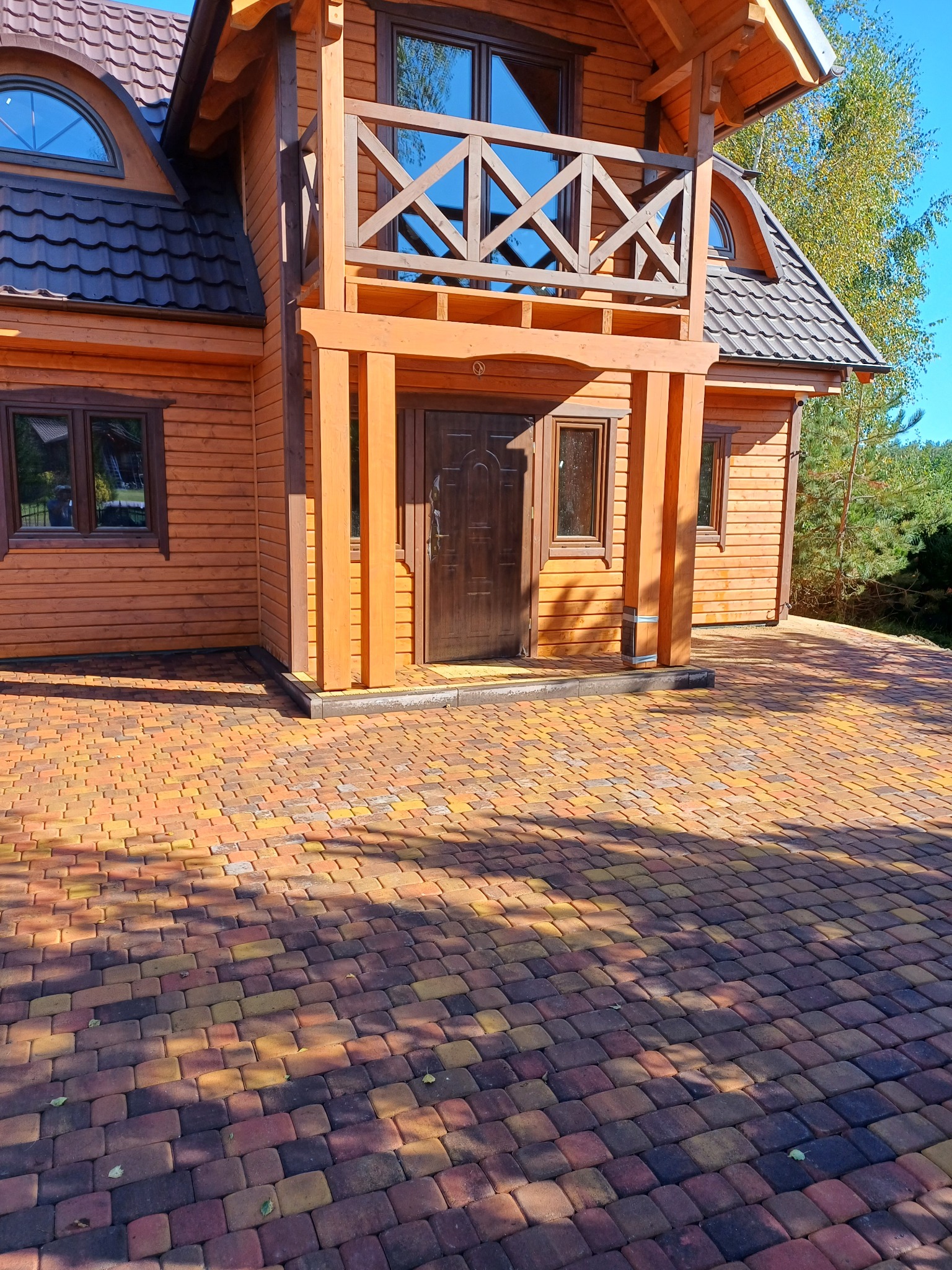 Drewniany dom z brązowymi oknami i balkonem, z wyłożonym kolorową kostką brukową podjazdem.