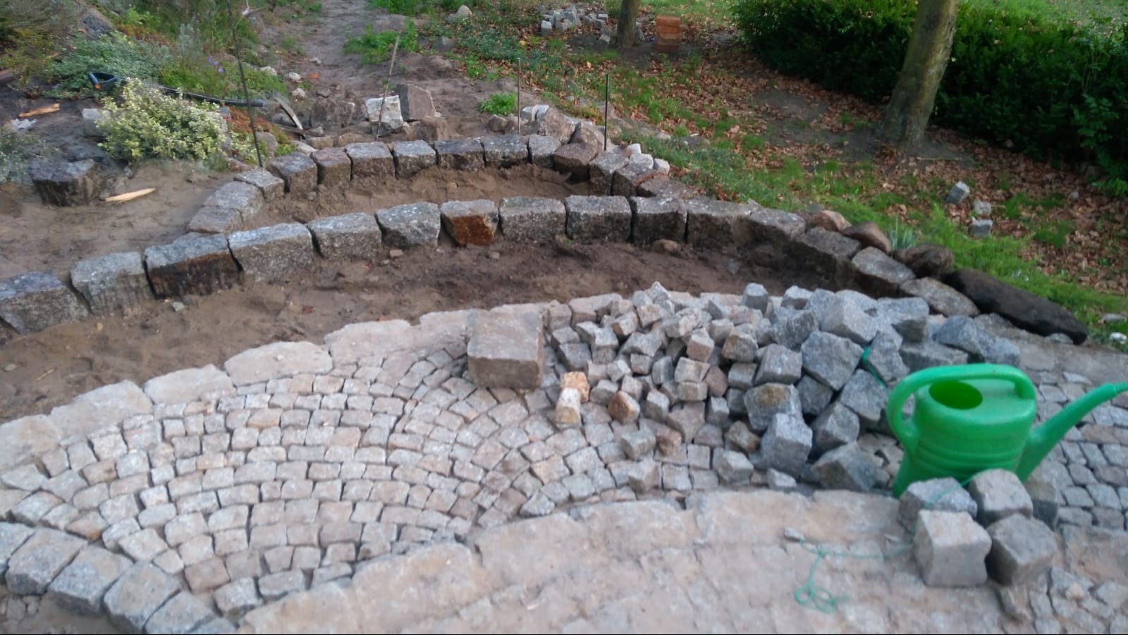 Układanie kamiennej ścieżki w ogrodzie, widoczne obrzeża z kostki granitowej, stos kamieni do dalszej pracy i zielona konewka.