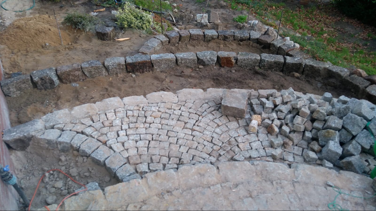 Fragment budowy tarasu z kamiennej kostki, widoczne ułożone fragmenty w kształcie łuku, obrzeża z większych kamieni i sterta kostki brukowej czekająca na ułożenie.