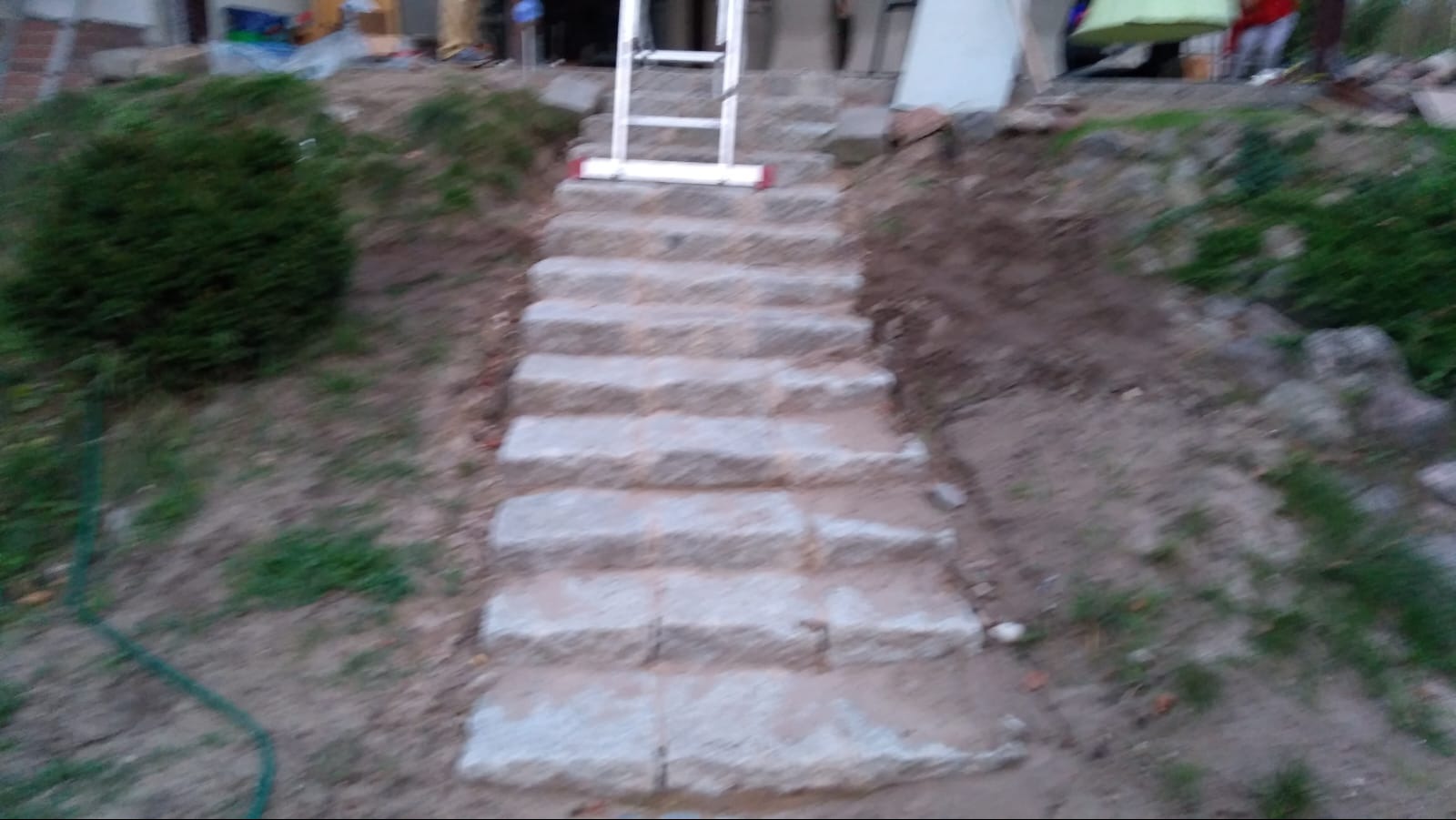 Kamienne schody zewnętrzne w trakcie budowy, widoczna drabina oparta o stopnie, ziemia i roślinność w tle.