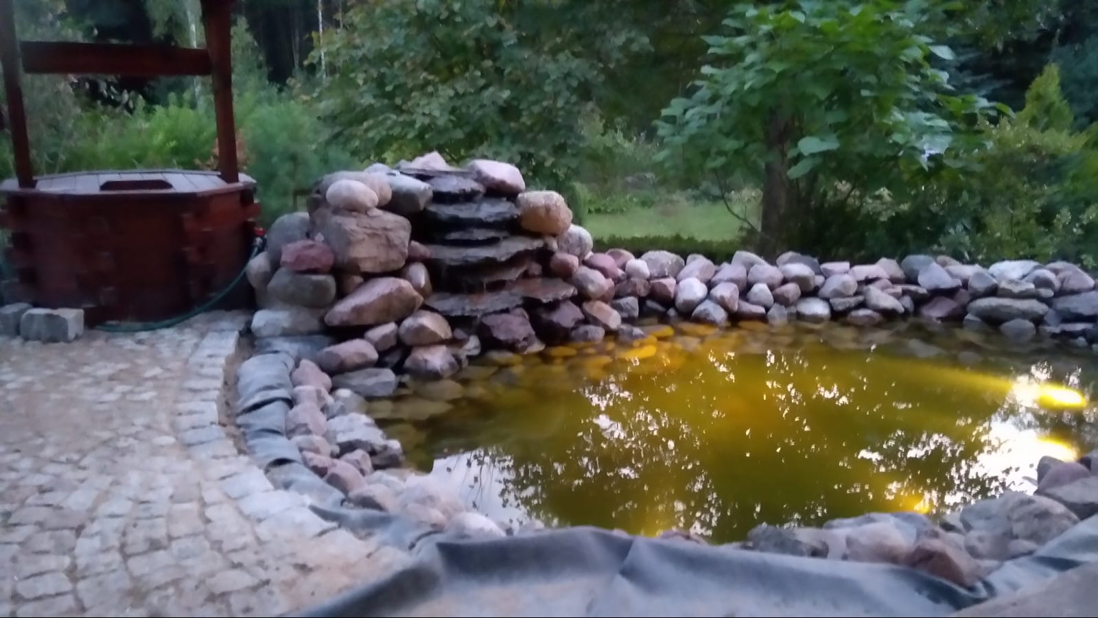 Oczko wodne z kamienną kaskadą i studnią z drewnianą obudową, otoczone brukowaną ścieżką i zielenią ogrodu.