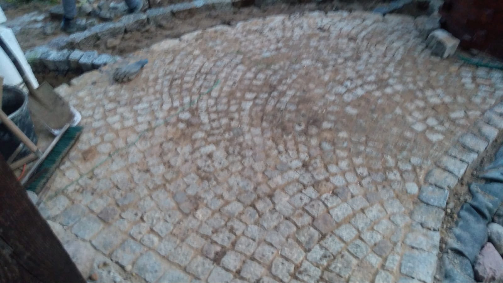 Niedokończona nawierzchnia z kostki granitowej ułożonej w łukowaty wzór, widoczne narzędzia murarskie i fragmenty niezabudowanego terenu.