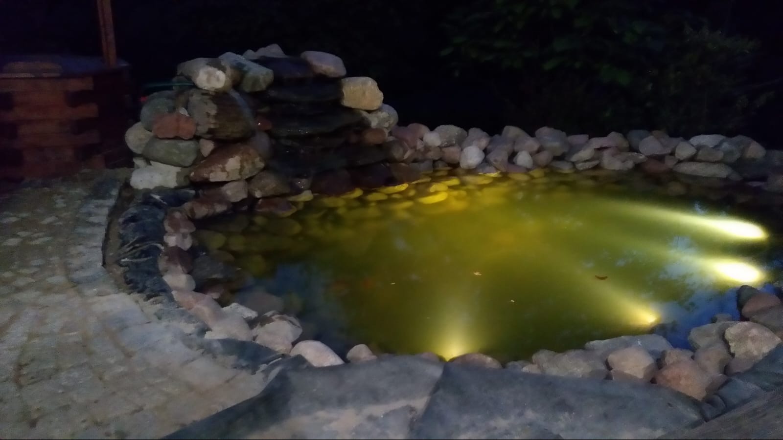 Oświetlony oczko wodne z kamienną kaskadą i obrzeżem z kostki, widoczne w nocy.