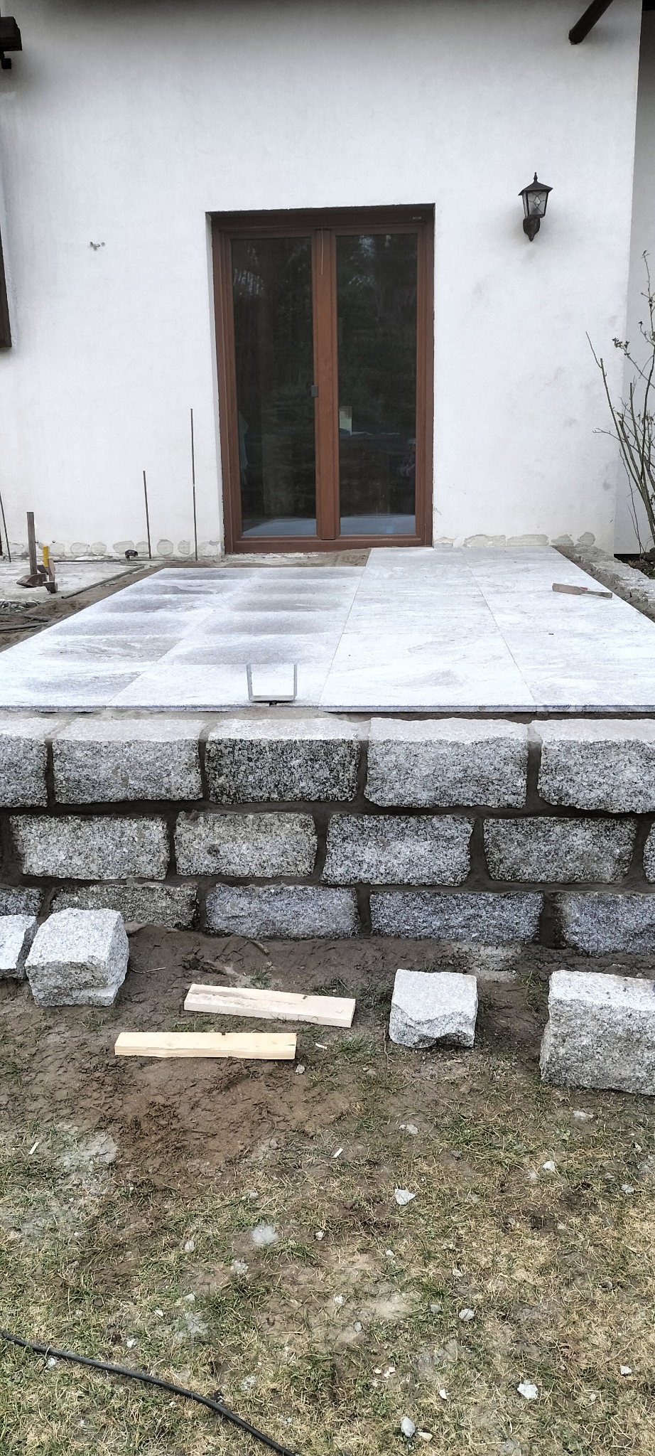 Wykonywanie tarasu z kamiennych płyt przy białym budynku, widoczne kamienne obrzeże, płyty tarasowe i narzędzia, etap prac w toku.