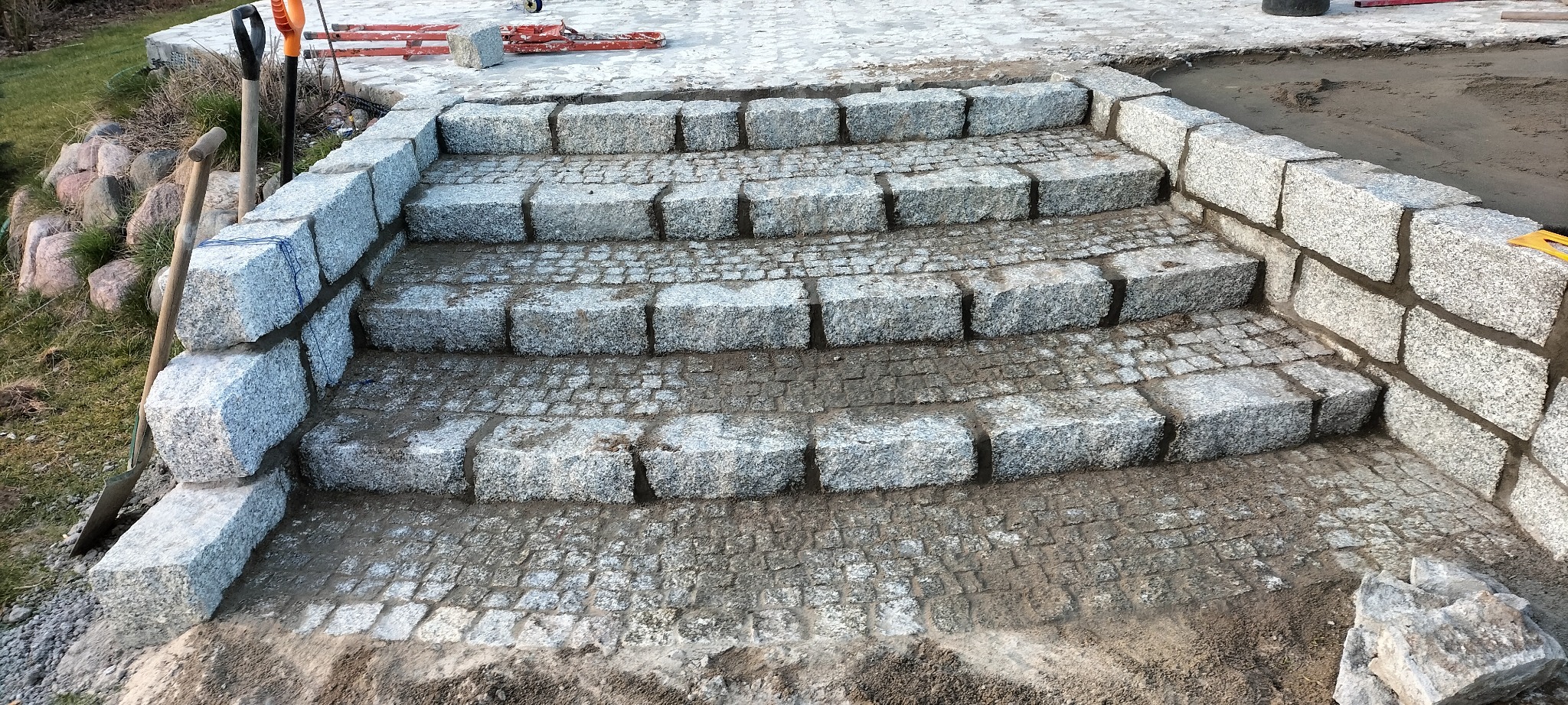 Schody z kostki granitowej w trakcie budowy, widoczne narzędzia i materiały budowlane, trawa w tle.
