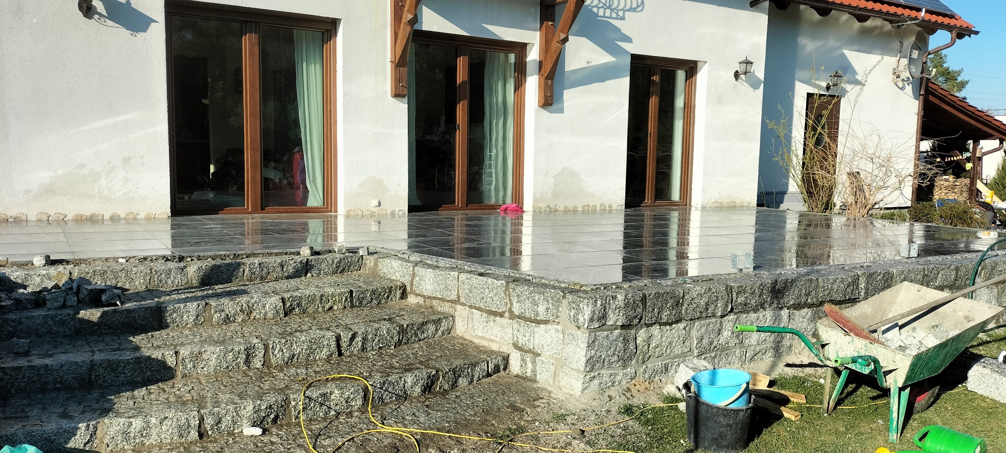 Granitowe schody prowadzące na taras z ciemnych płytek, widoczne narzędzia budowlane i taczka.