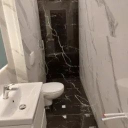 Nowoczesna łazienka z białymi i czarnymi płytkami imitującymi marmur, umywalką na szafce i toaletą. Widoczne chromowane elementy armatury i wystające rury z oznaczeniami kolorystycznymi.