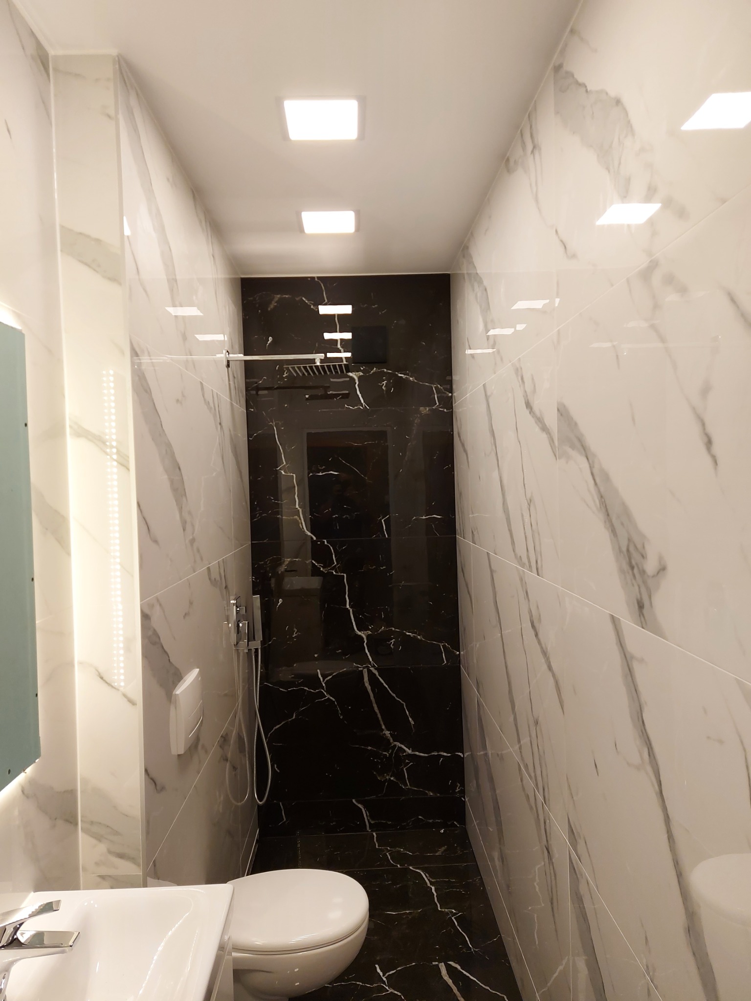 Wąska łazienka z kabiną prysznicową typu walk-in wyłożoną czarnymi płytkami imitującymi marmur, kontrastującymi z białymi płytkami z szarymi żyłami na pozostałych ścianach, podświetlona...