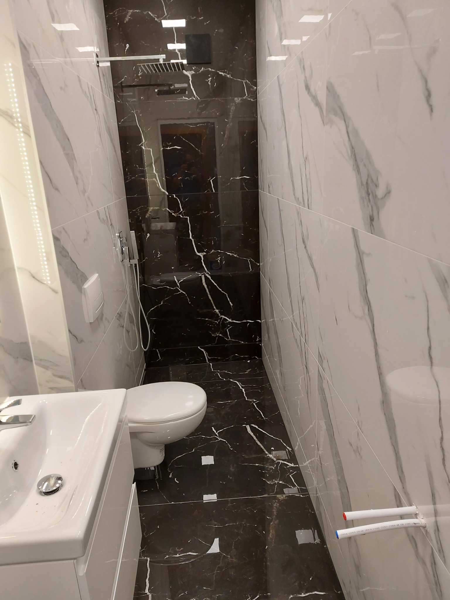 Wąska łazienka z białymi i czarnymi marmurkowymi płytkami, biała umywalka z szafką i toaleta, nowoczesna deszczownica, widoczne rury doprowadzające wodę.