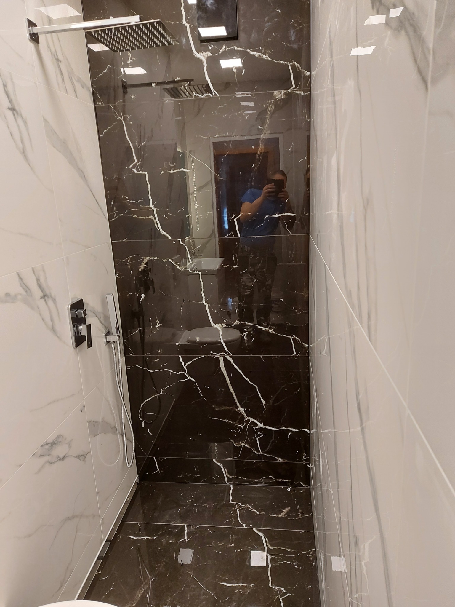 Wnętrze kabiny prysznicowej wyłożonej płytkami imitującymi marmur w odcieniach bieli i brązu z czarnymi żyłami, deszczownica zamontowana na suficie, widoczne odbicie fotografa w szklanej ściance.