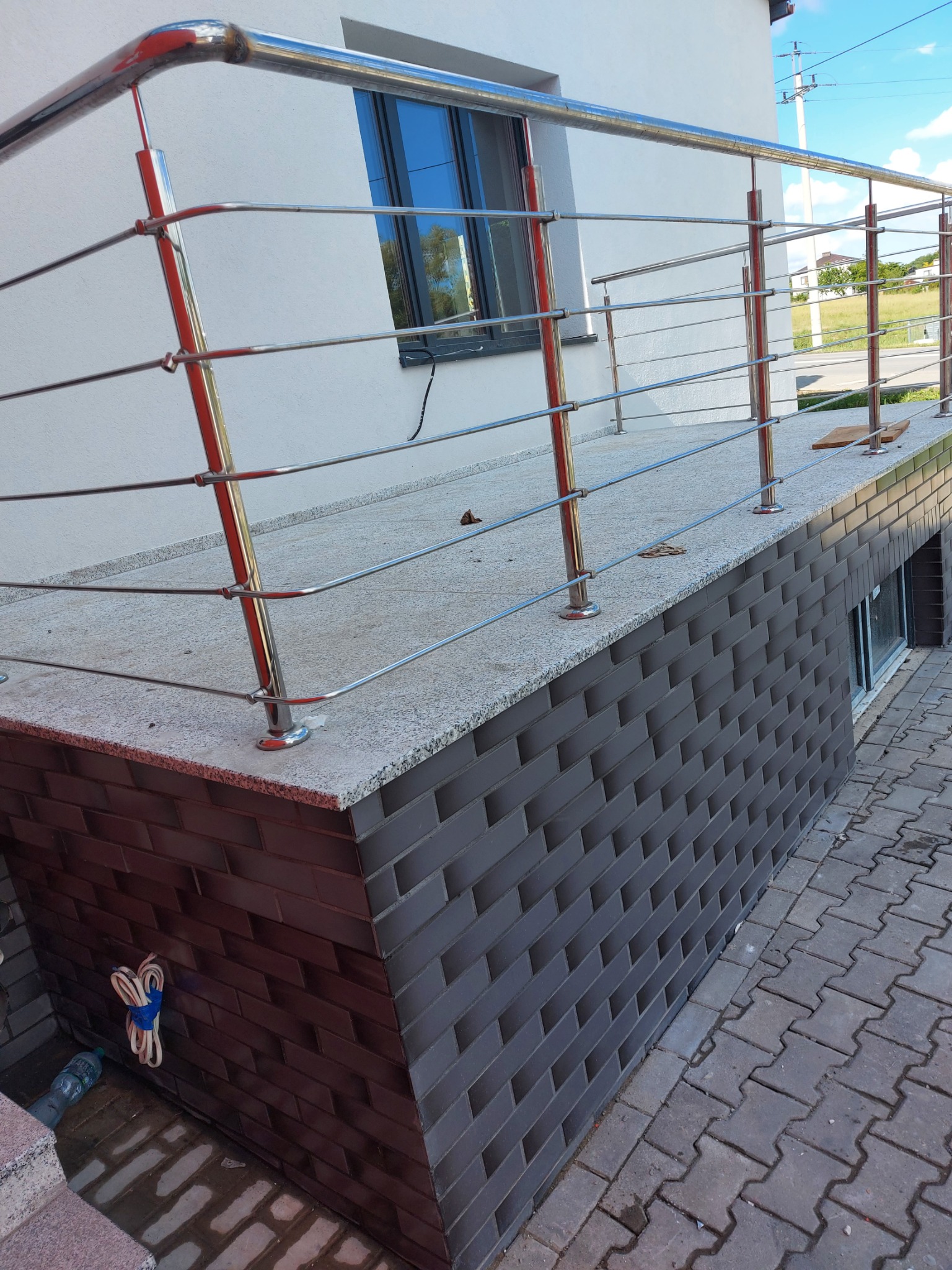 Nowoczesna balustrada ze stali nierdzewnej na tarasie wykończonym cegłą klinkierową i szarym kamieniem, widoczne okno piwniczne i kostka brukowa.