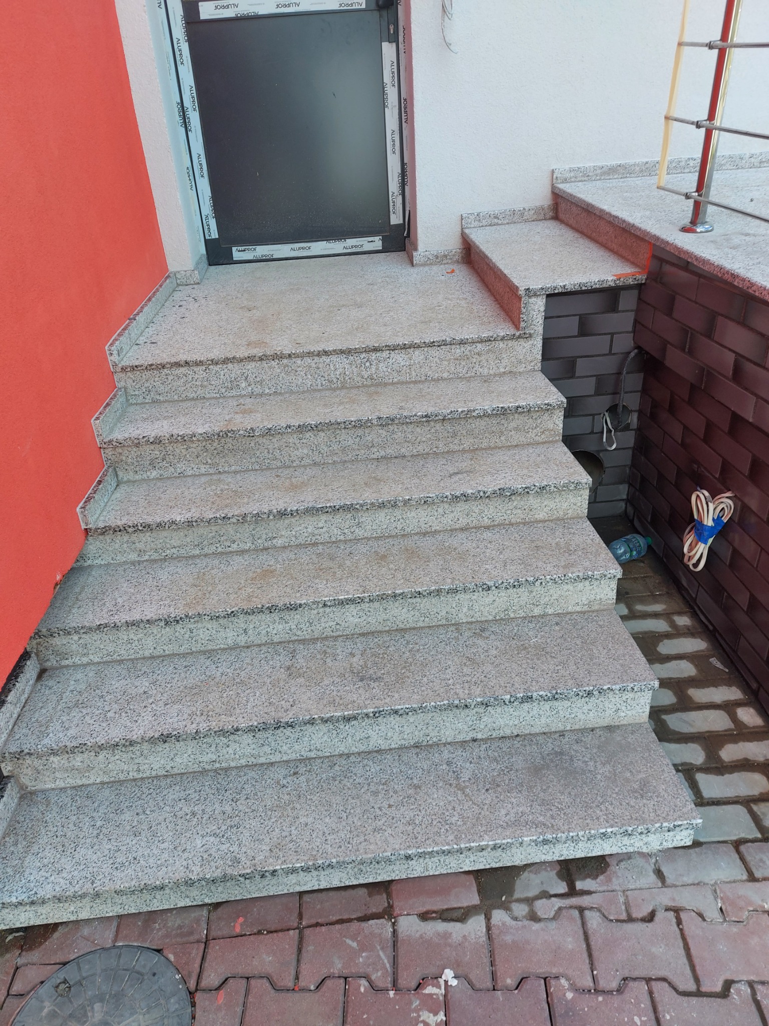 Schody zewnętrzne z jasnego granitu prowadzące do drzwi wejściowych, z widocznymi elementami wykończenia elewacji i kostki brukowej.