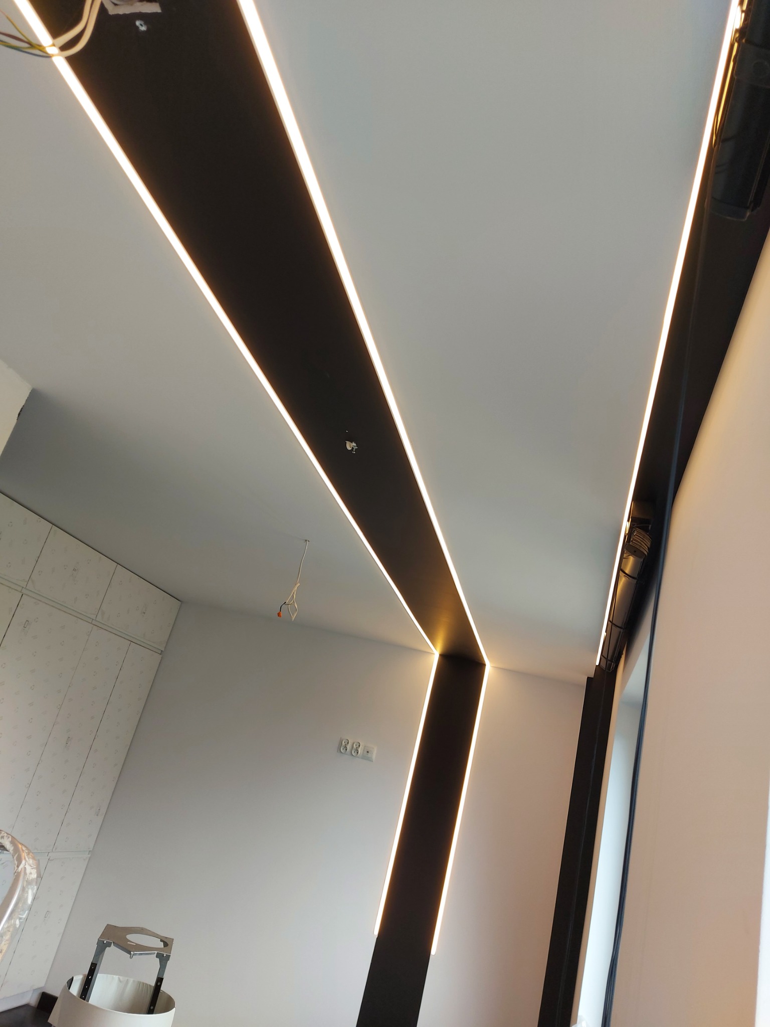 Nowoczesne oświetlenie sufitowe z podłużnymi lampami LED w czarnych panelach, widok z dołu, fragment ściany z szafą w tle, widoczne kable i gniazdka elektryczne.