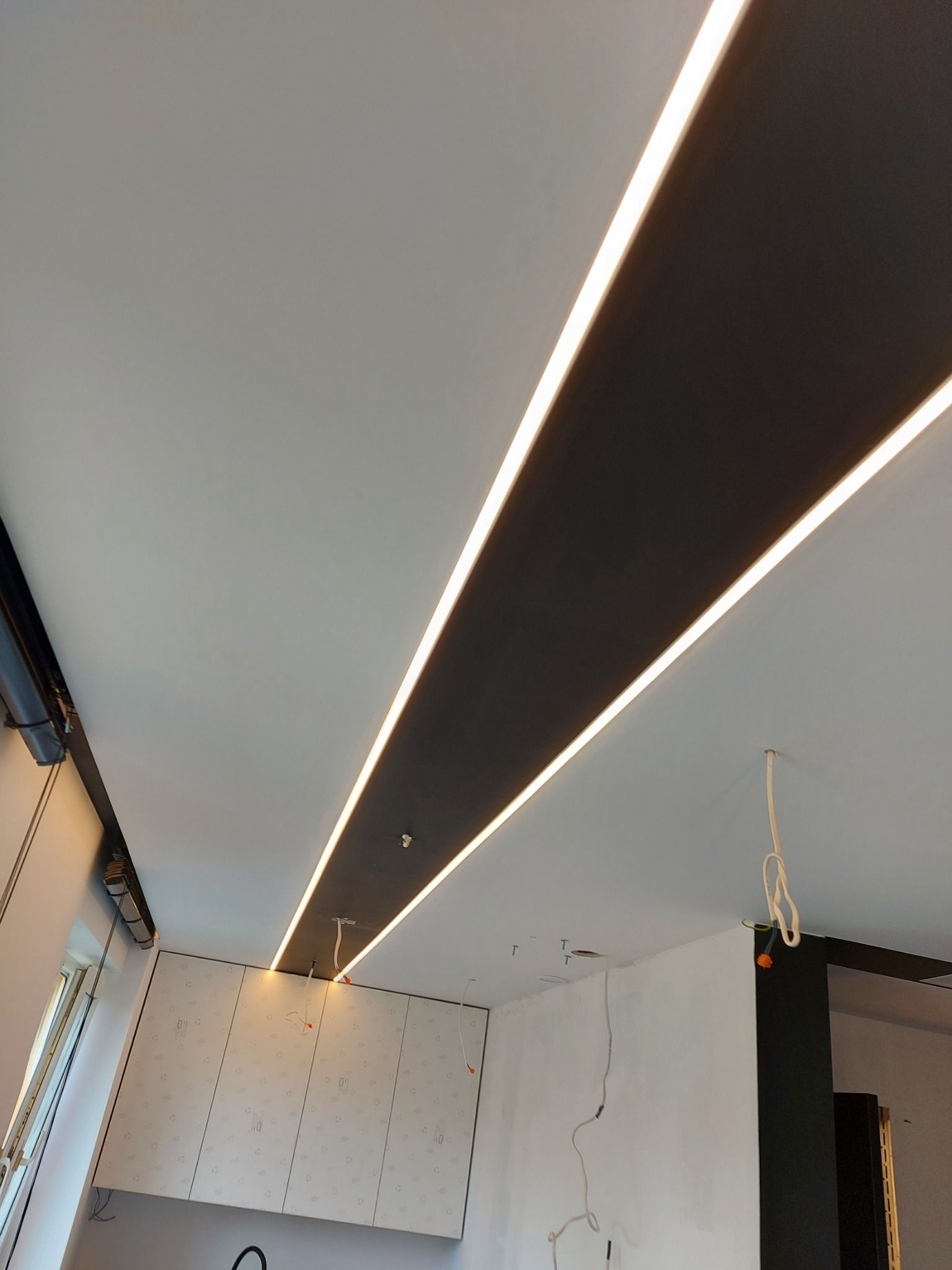 Nowoczesny sufit z podwójnym pasem czerni i liniowym oświetleniem LED, widoczne niedokończone instalacje elektryczne i fragment ściany z szafkami.