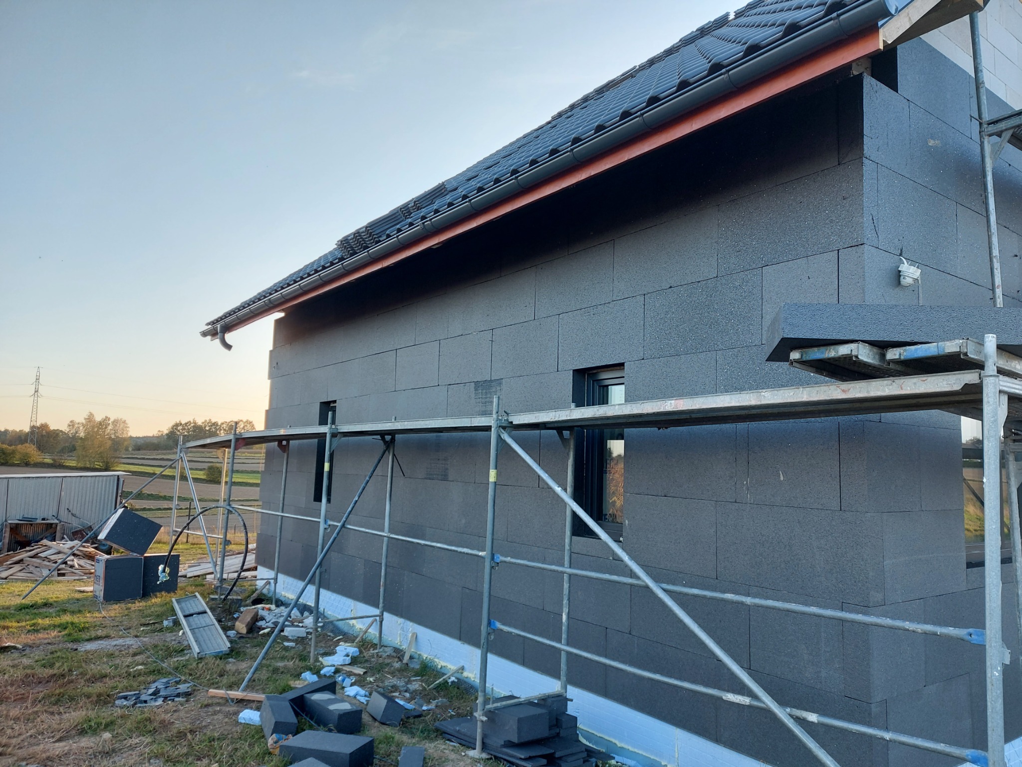 Dom jednorodzinny w trakcie ocieplania elewacji styropianem grafitowym, widoczne rusztowanie, fragment dachu z rynną i niezabezpieczone okno.