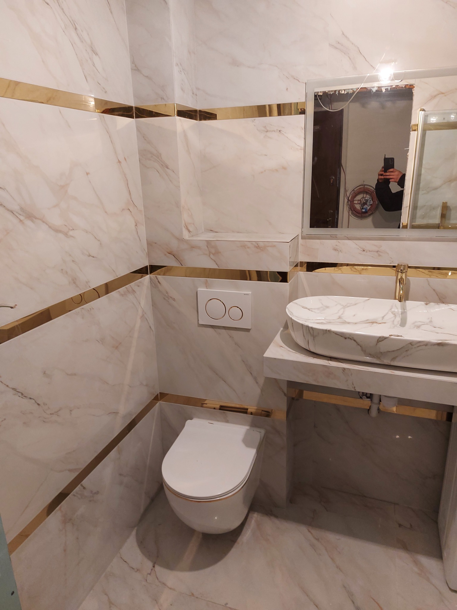 Nowoczesna łazienka z marmurowymi płytkami, złotymi akcentami i owalną umywalką nablatową. W lustrze odbija się osoba robiąca zdjęcie.