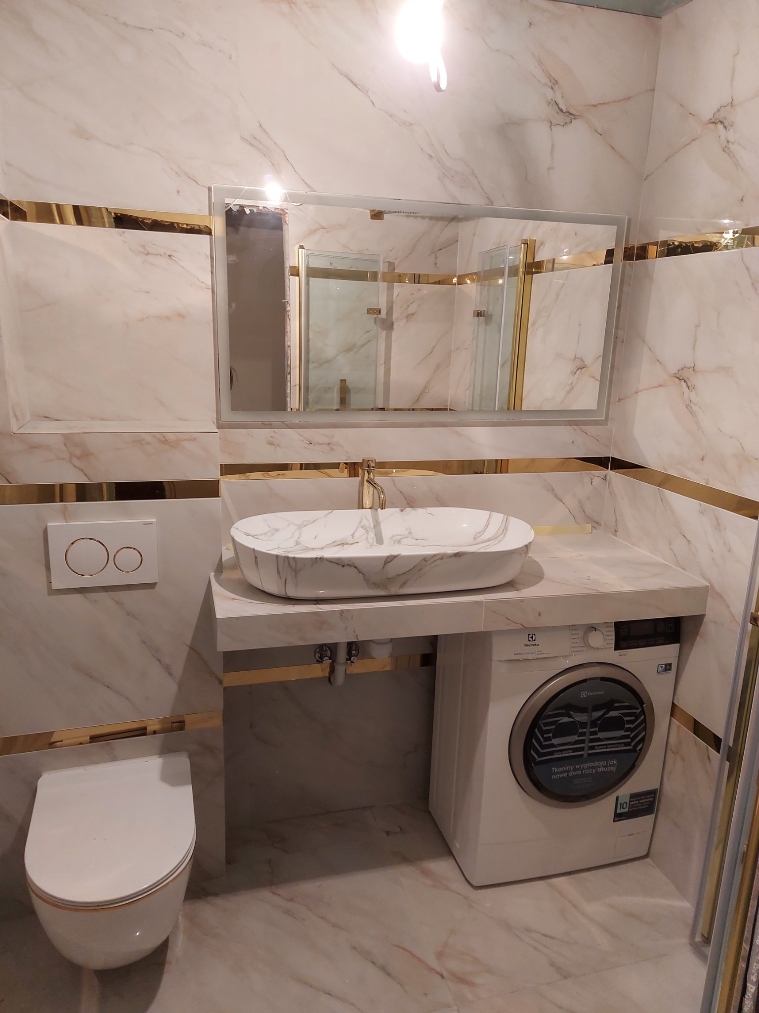 Elegancka łazienka z marmurowymi płytkami, złotymi akcentami, umywalką nablatową w marmurowy wzór, podwieszaną toaletą, lustrem z podświetleniem i pralką Electrolux pod blatem.