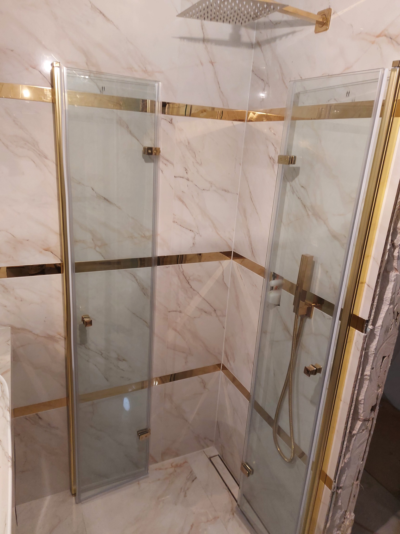 Kabina prysznicowa narożna z dwuskrzydłowymi drzwiami, wykończona złotymi profilami i armaturą, na tle marmurowych płytek z złotymi pasami dekoracyjnymi, widoczny odpływ liniowy.