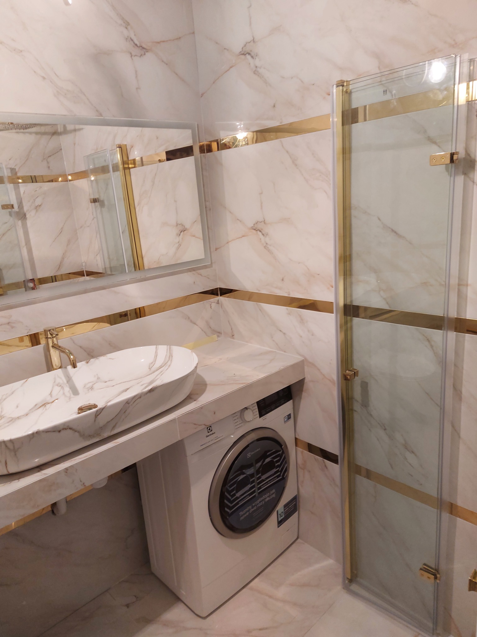 Elegancka łazienka z marmurowymi płytkami, owalną umywalką nablatową, złotą armaturą i kabiną prysznicową ze złotymi profilami. Pod blatem umywalkowym pralka Electrolux.