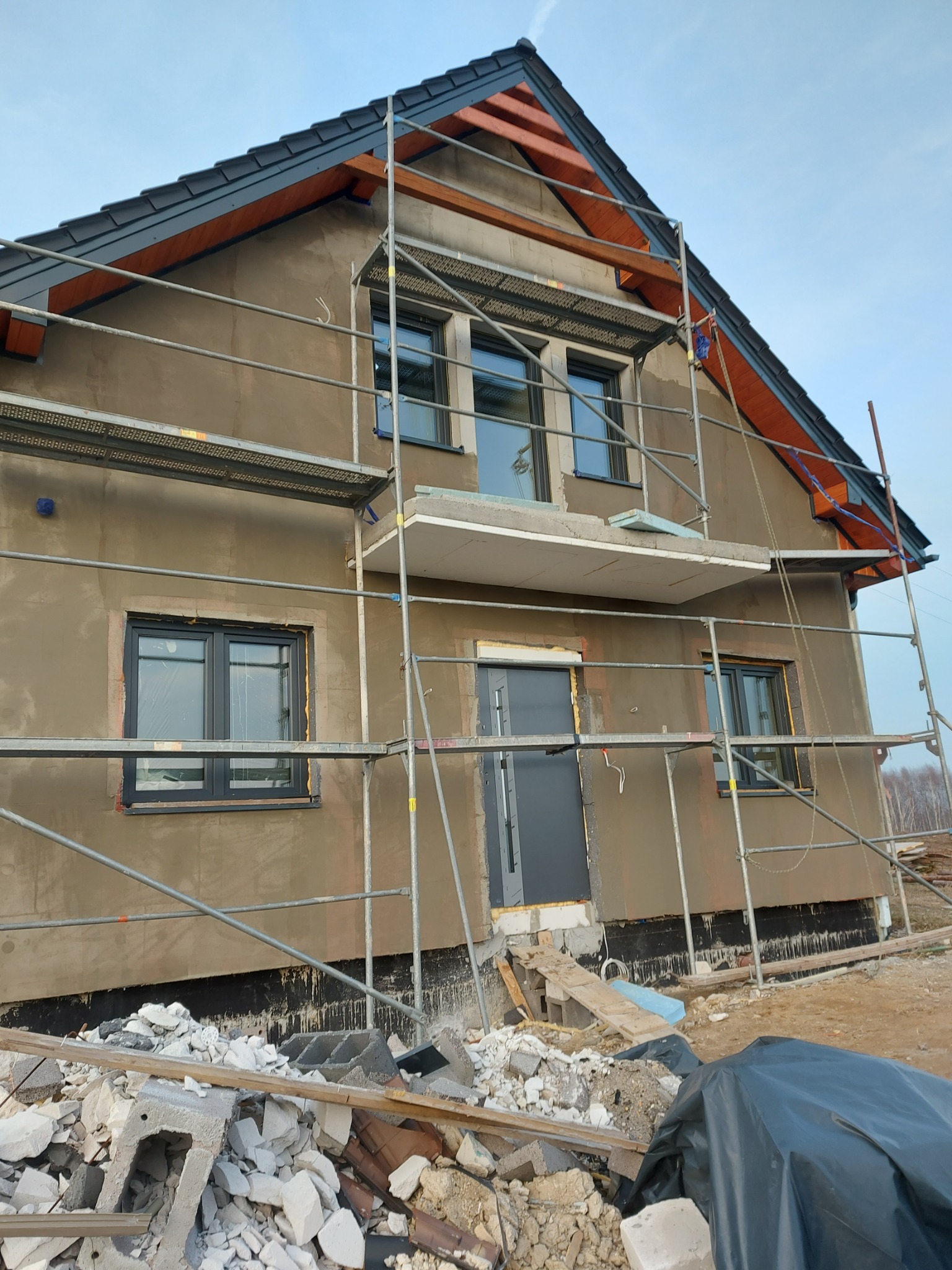 Dom jednorodzinny w trakcie prac elewacyjnych: szare okna, otynkowana ściana, rusztowanie i gruz na ziemi.