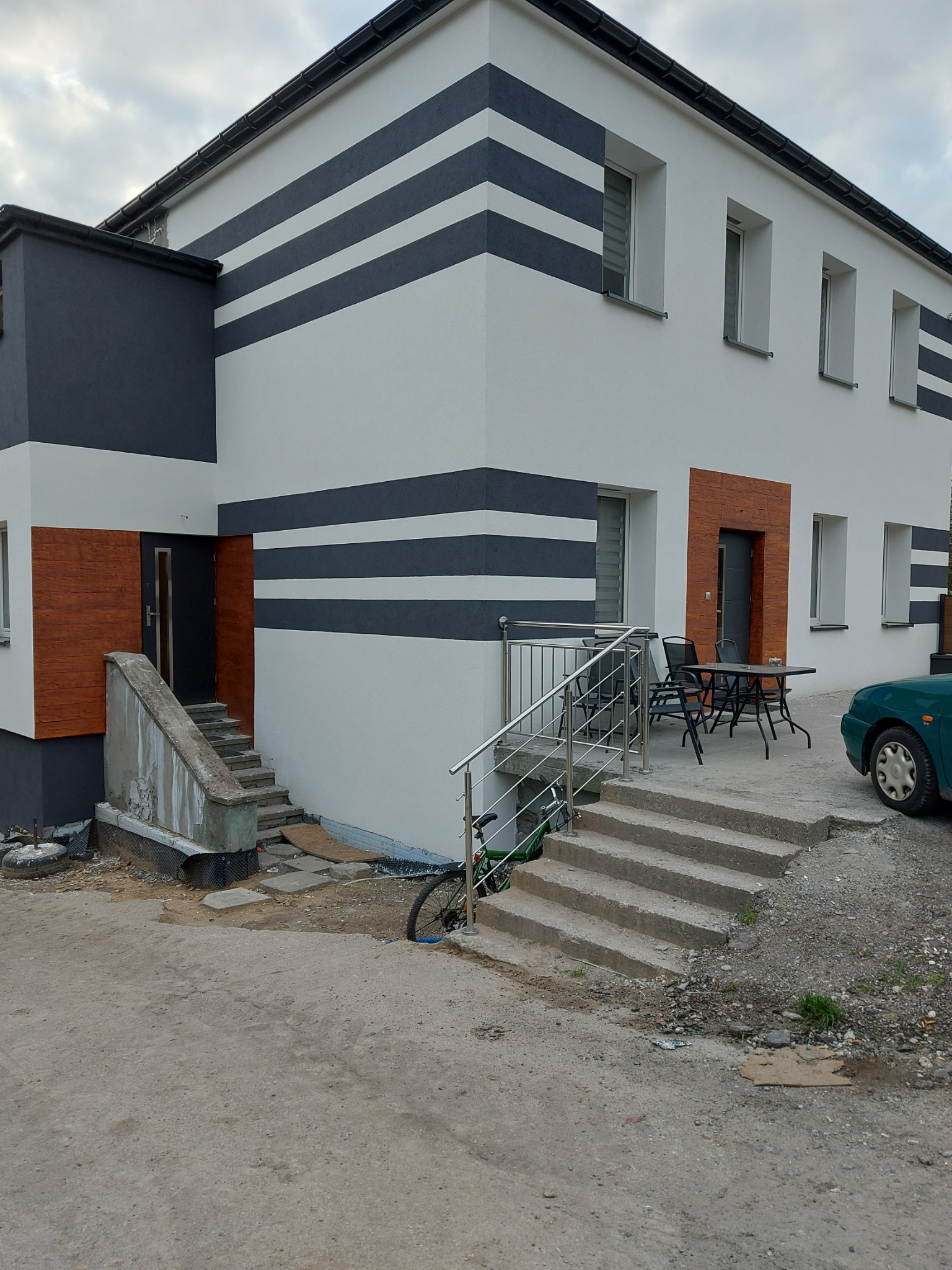 Dwukondygnacyjny dom z białą elewacją, ozdobiony poziomymi pasami w kolorze grafitowym, z elementami dekoracyjnymi imitującymi drewno wokół drzwi wejściowych, widoczne schody wejściowe z metalową...