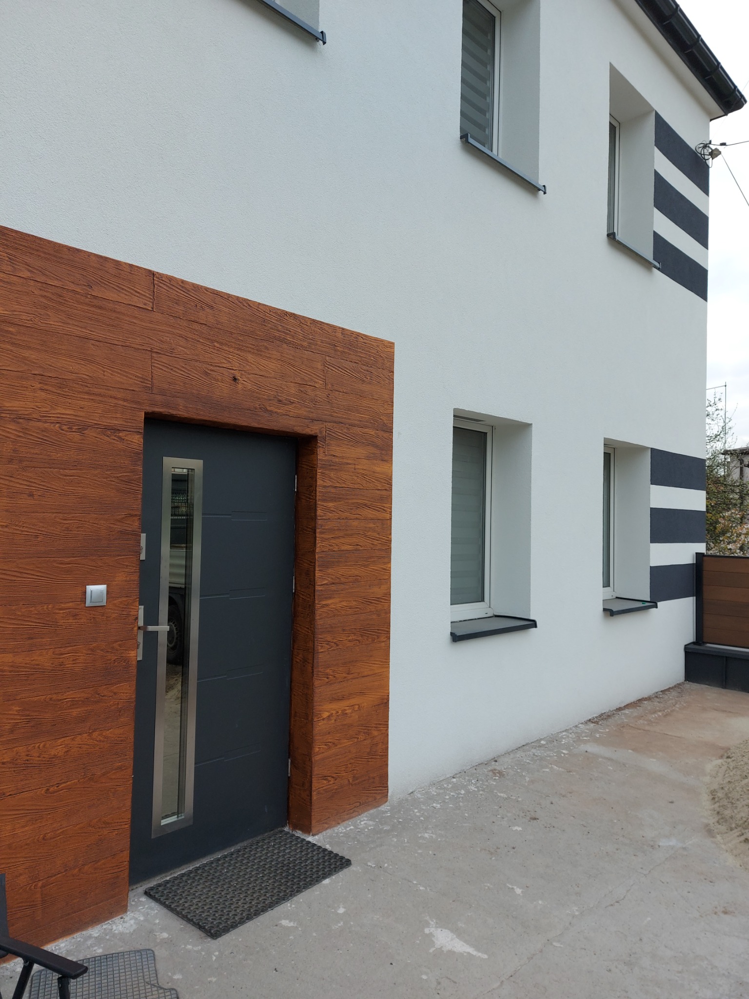 Nowoczesny dom z białą elewacją, drewnopodobną okładziną wokół wejścia i antracytowymi akcentami w postaci pasów i drzwi.