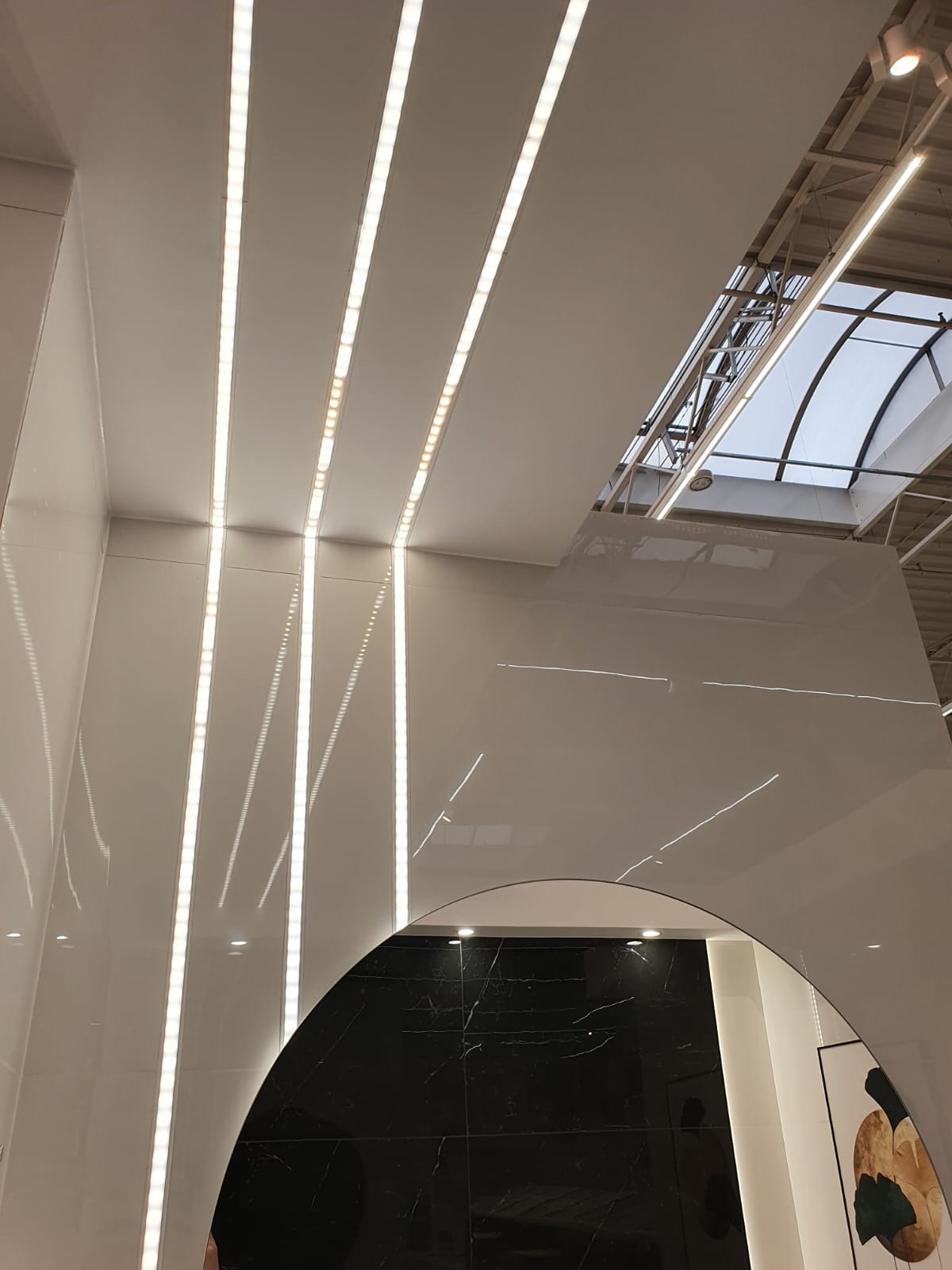 Białe panele sufitowe z wbudowanym oświetleniem LED, widok z dołu, odbicie czarnej marmurkowej ściany w zaokrąglonym lustrze, fragment okna dachowego w tle.