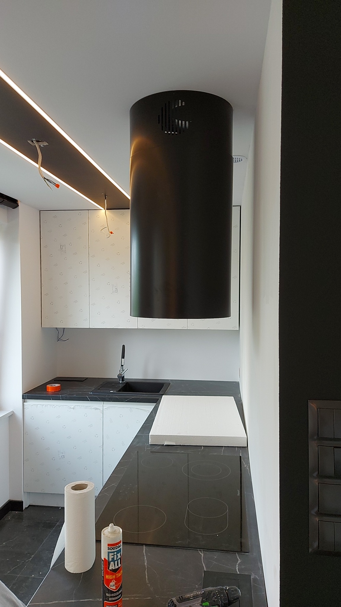 Wnętrze kuchni w trakcie montażu: czarny, cylindryczny okap nad płytą indukcyjną, szafki z folią ochronną, widoczny klej montażowy Soudal Fix All i narzędzia.