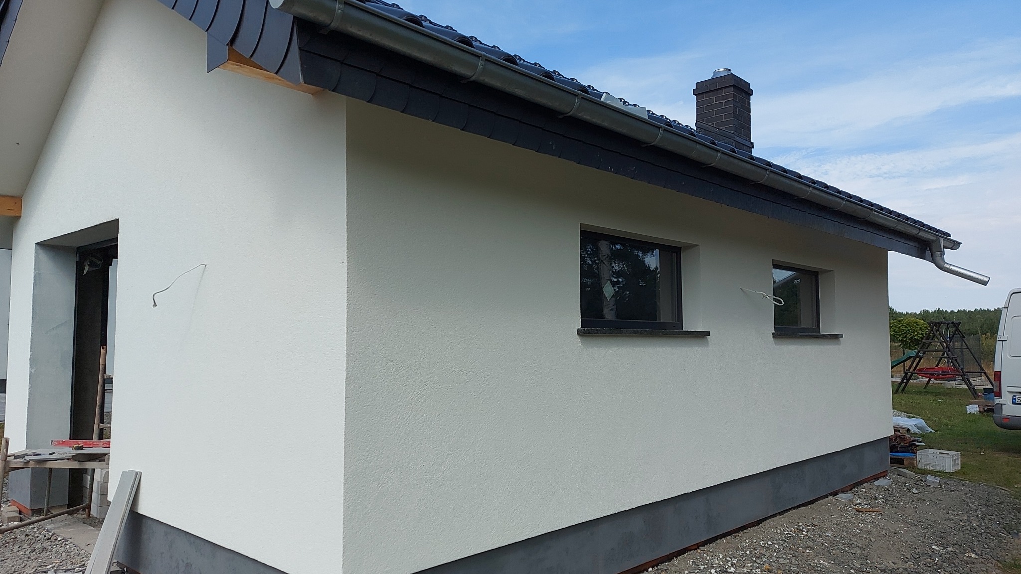 Świeżo pomalowana elewacja domu jednorodzinnego w jasnym odcieniu, widoczne detale wykończeniowe, takie jak czarna podmurówka, okna z ciemnymi ramami i dachówka, na elewacji widoczny wystający...