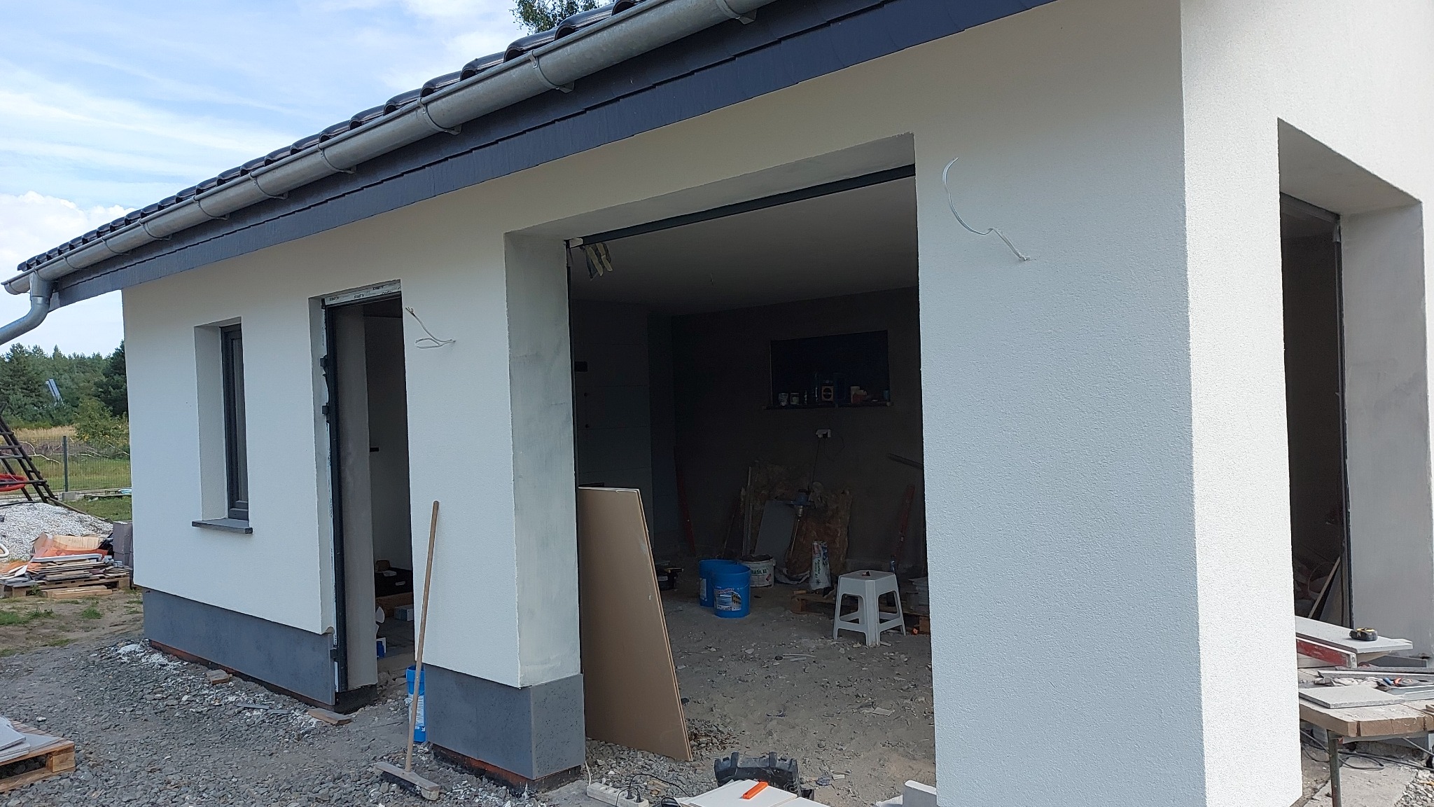 Nowo wykończony budynek gospodarczy z białymi ścianami i ciemnoszarą podmurówką w trakcie wykańczania, widoczne narzędzia i materiały budowlane wewnątrz i na zewnątrz budynku.