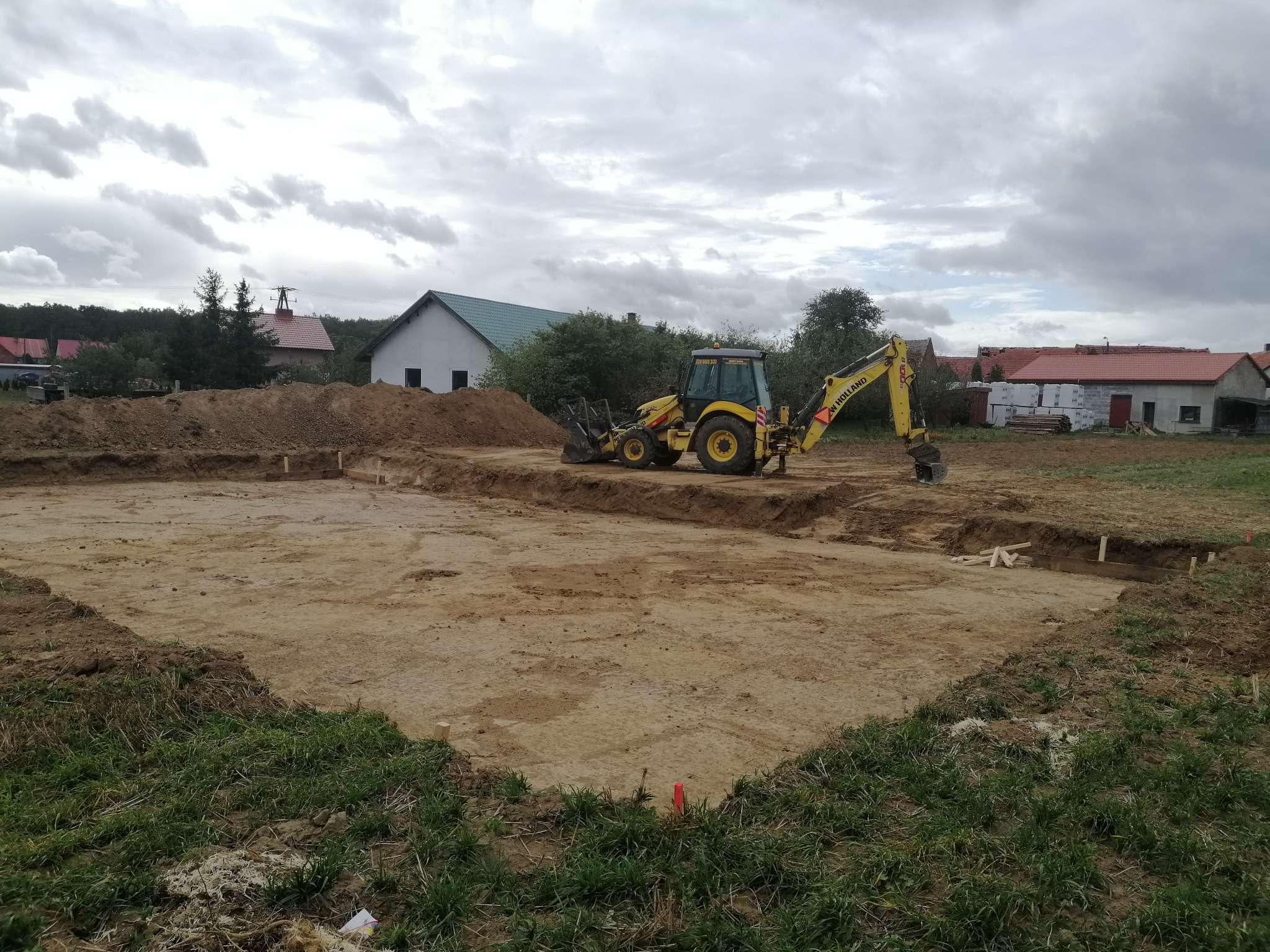 Żółta koparko-ładowarka New Holland podczas wykopu pod fundamenty domu jednorodzinnego na działce z widokiem na wiejską zabudowę w pochmurny dzień.