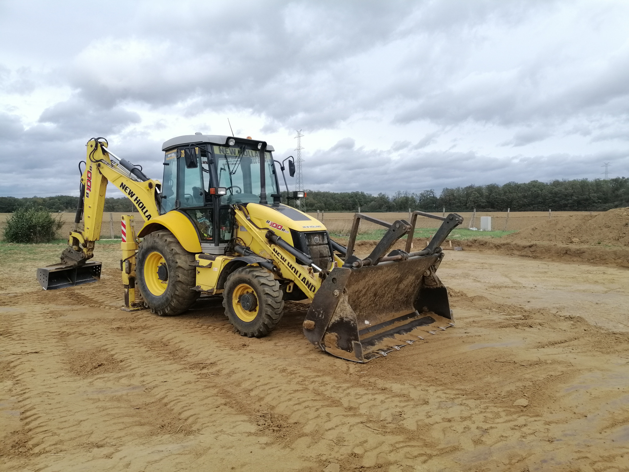 Żółta koparko-ładowarka New Holland B100B na piaszczystym terenie budowy z wykopem w tle, pochmurne niebo.