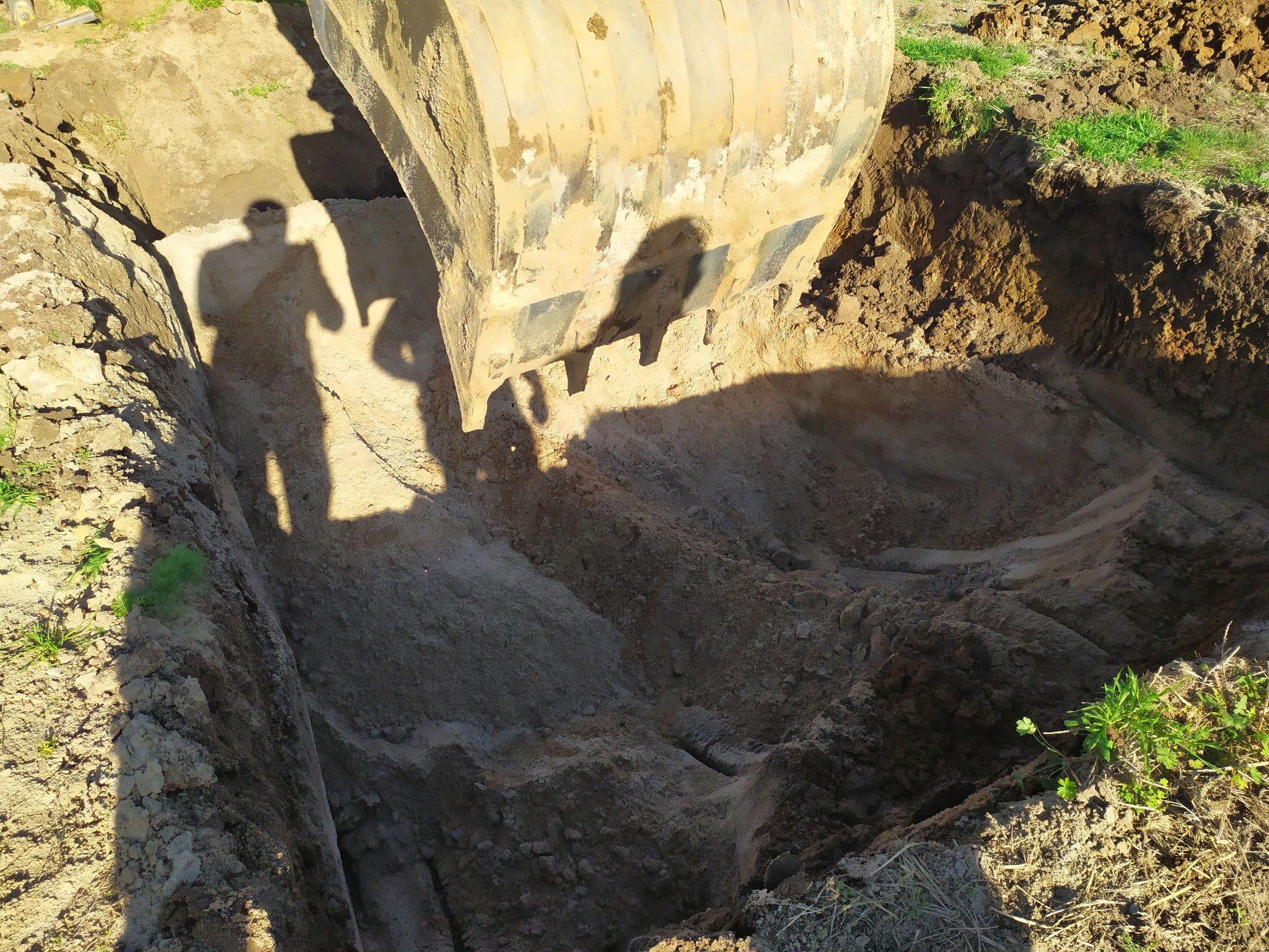 Głęboki wykop w ziemi, z cieniem koparki i operatora widocznym na ścianie, przygotowany pod instalację szamba betonowego.