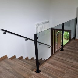 HardGlass Arkadiusz Król - Wewnętrzne schody wykończone płytkami imitującymi drewno, zabezpieczone nowoczesną balustradą ze szkła i czarnych, metalowych słupków, z widocznym grzejnikiem na ścianie i drzwiami wejściowymi w tle.