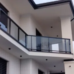 Nowoczesna szklana balustrada balkonowa z czarnymi profilami, widok z dołu na elewację budynku z oświetleniem punktowym.