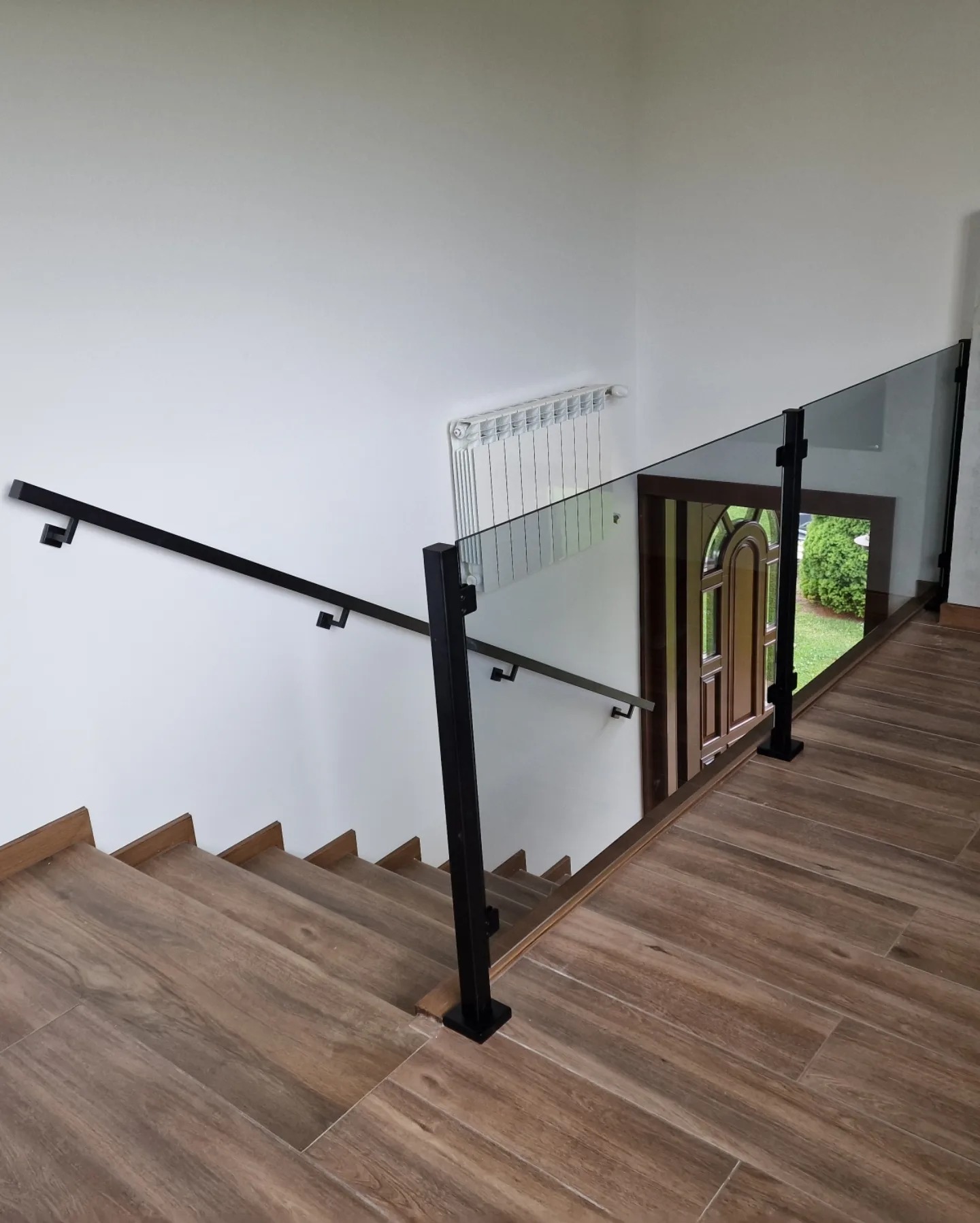 Wewnętrzne schody wykończone płytkami imitującymi drewno, zabezpieczone nowoczesną balustradą ze szkła i czarnych, metalowych słupków, z widocznym grzejnikiem na ścianie i drzwiami wejściowymi w tle.