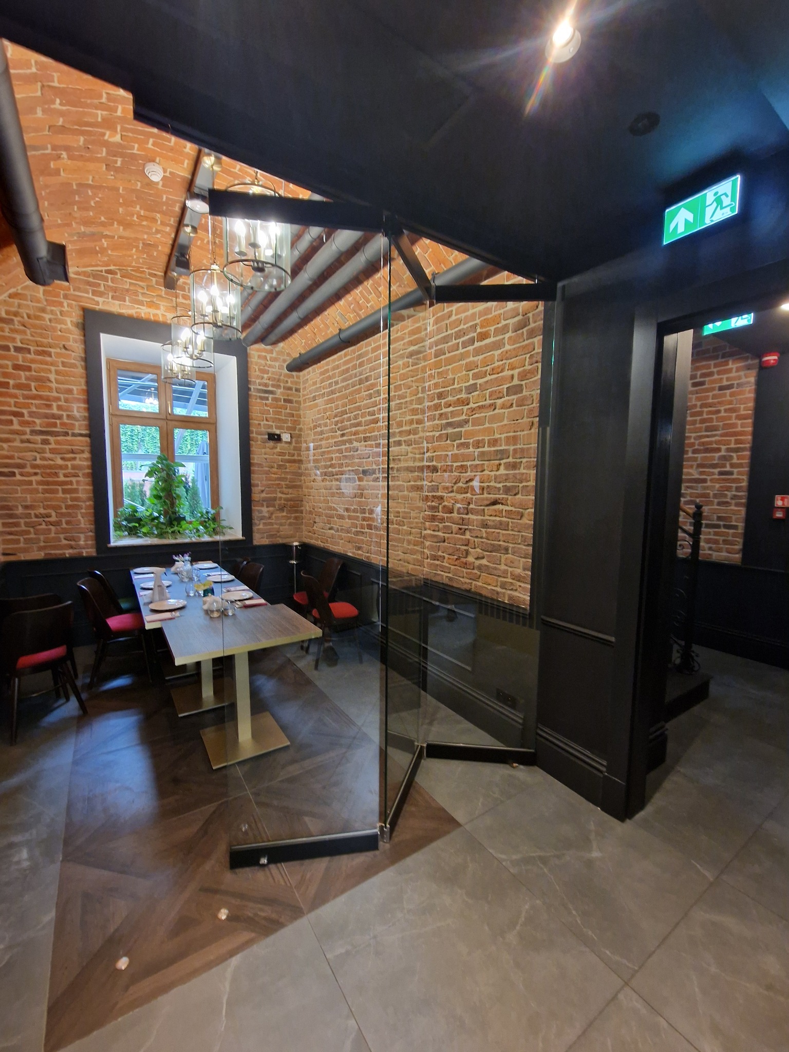 Szklane drzwi wahadłowe oddzielające część restauracji z ceglanymi ścianami i nakrytym stołem, widoczny znak ewakuacyjny.
