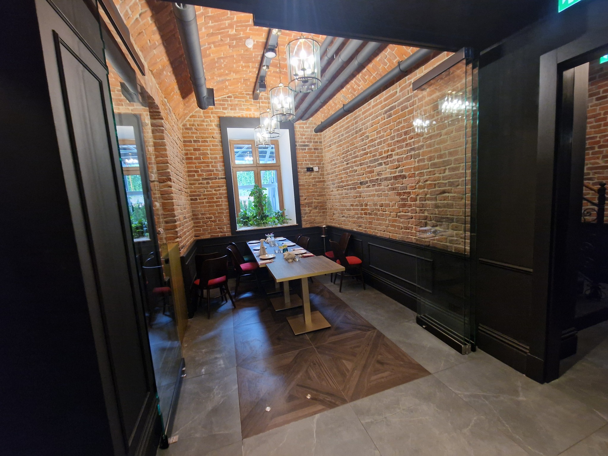 Szklane drzwi oddzielające salę restauracyjną z ceglanymi ścianami i drewnianą podłogą od ciemniejszego pomieszczenia, widoczne nakryte stoły.