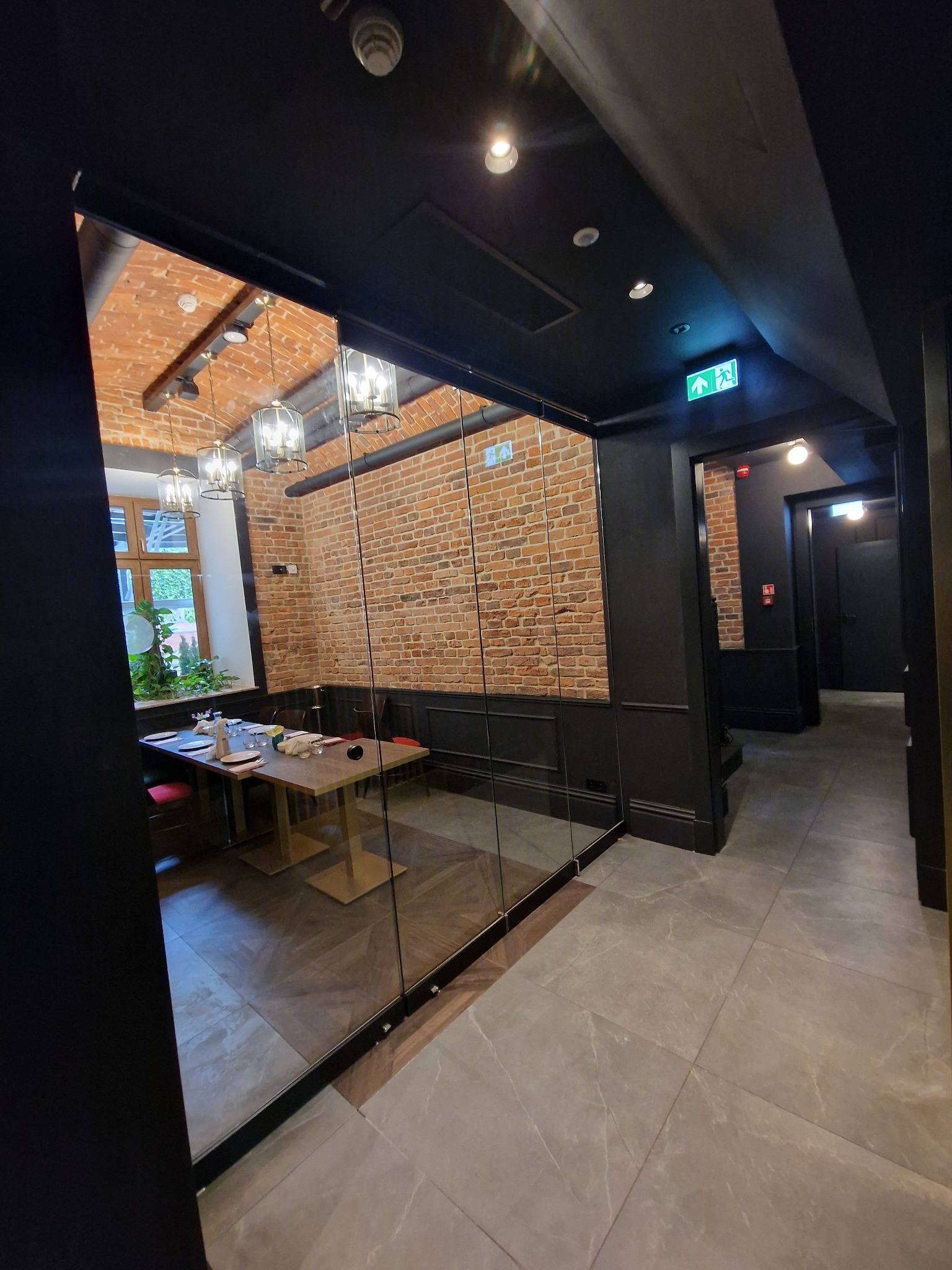 Szklana ścianka działowa oddzielająca salę restauracyjną z ceglaną ścianą i nakrytym stołem od korytarza z ciemnymi ścianami i podłogą z płytek.