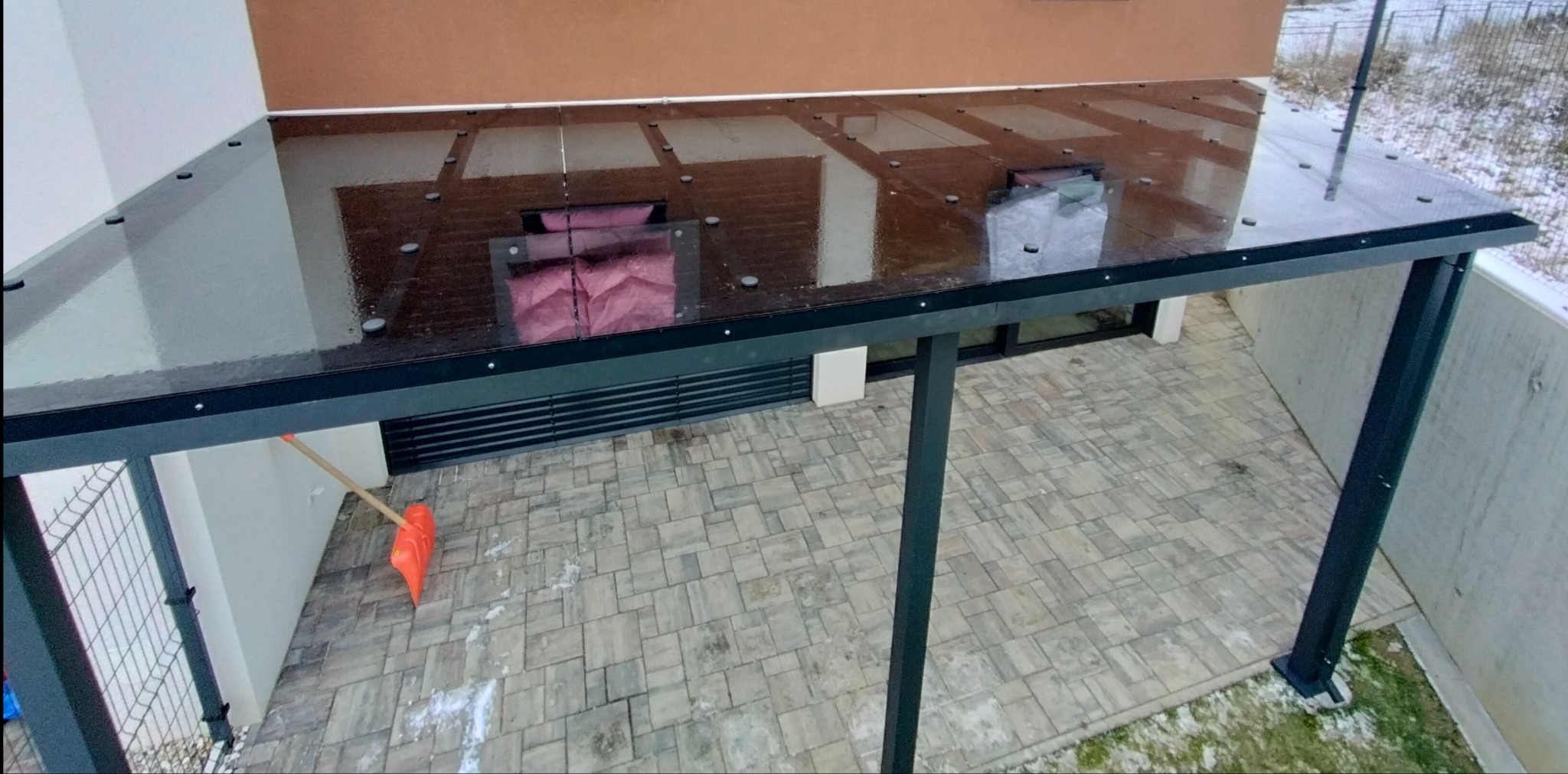 Nowoczesna pergola metalowa z przezroczystym szklanym dachem, widoczna z góry, z pomarańczową łopatą do śniegu opartą o ścianę w tle.