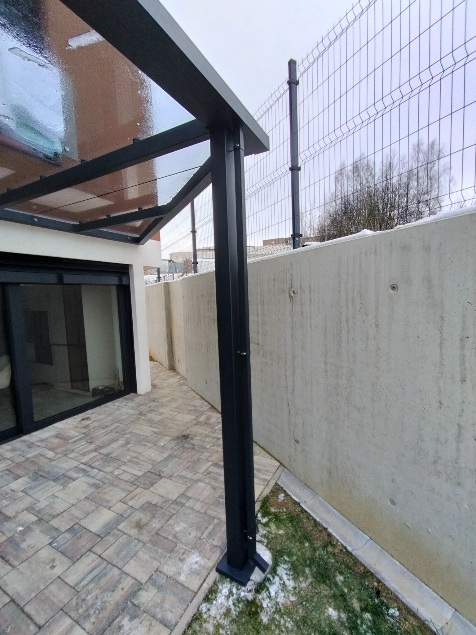 Nowoczesna pergola z metalową konstrukcją i przezroczystym zadaszeniem, przylegająca do budynku, z widoczną kostką brukową na tarasie i betonowym ogrodzeniem w tle.