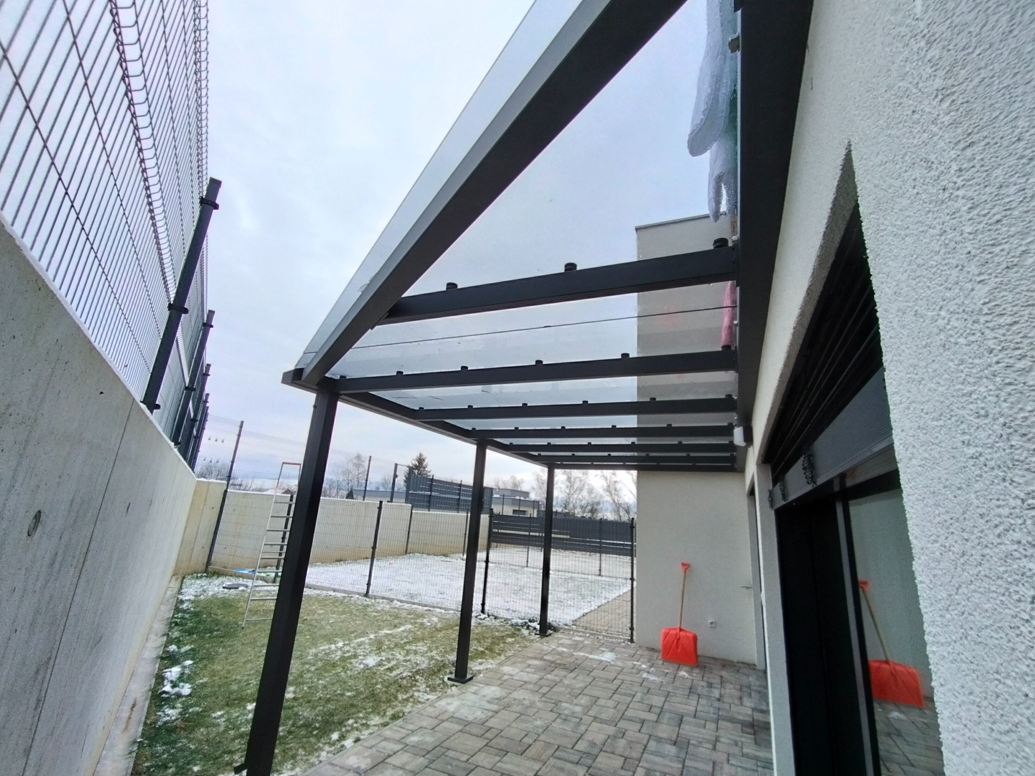 Nowoczesna pergola z metalową konstrukcją i szklanym dachem, widoczna z perspektywy pod nią, z elementami zimowego krajobrazu w tle i czerwoną łopatą do śniegu opartą o ścianę budynku.