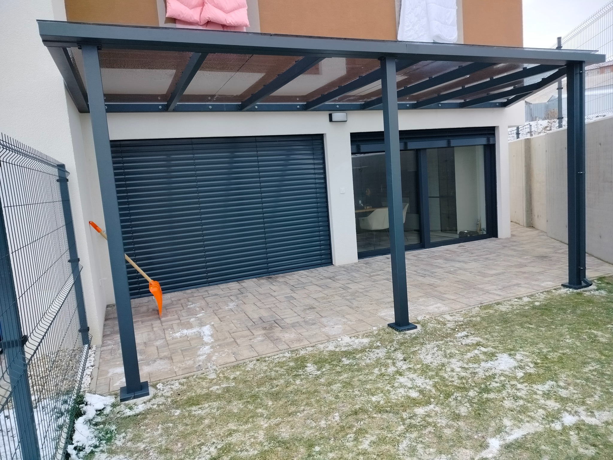 Metalowa pergola z przezroczystym zadaszeniem, przylegająca do budynku z zamkniętymi roletami i szklanymi drzwiami, z widoczną łopatą do śniegu i resztkami śniegu na trawniku.